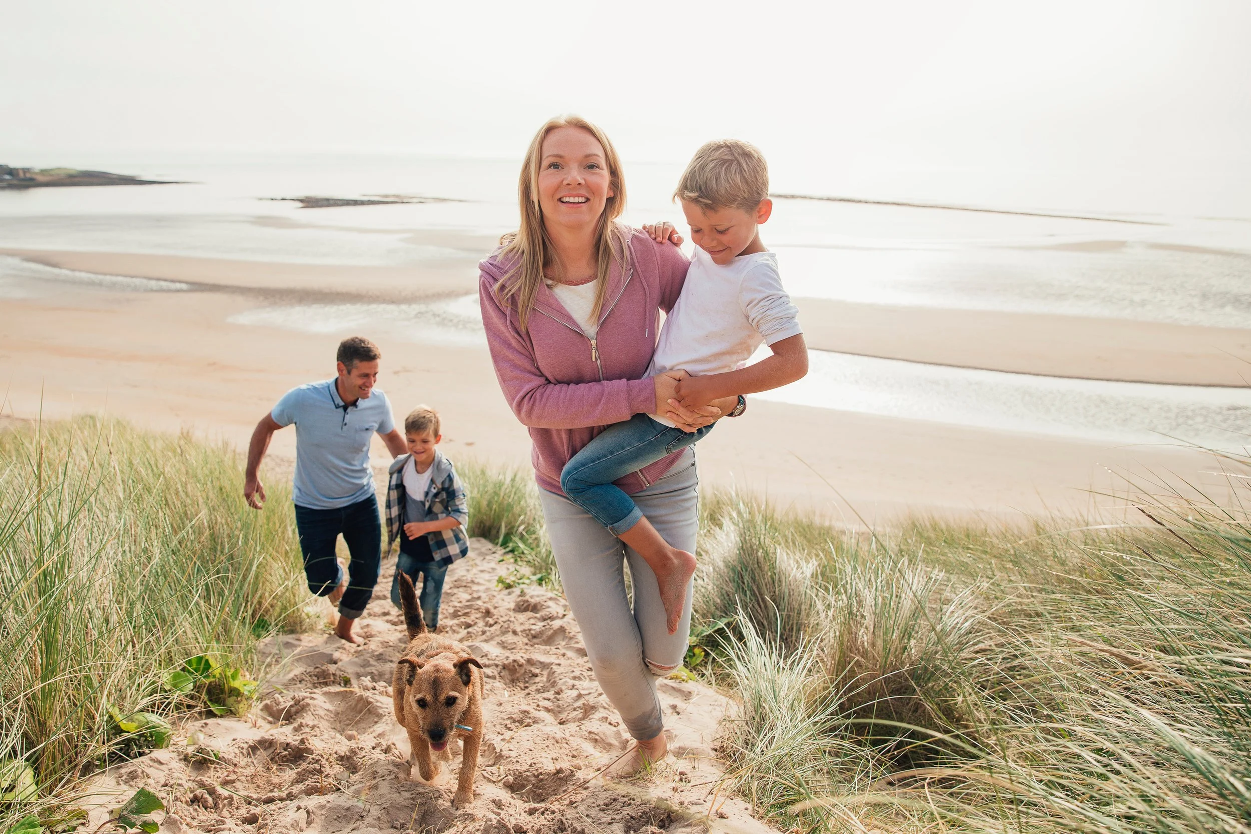 A smiling woman holding a young girl as they walk on a sandy beach with grass and a dog in the foreground and two men and a boy in the background.