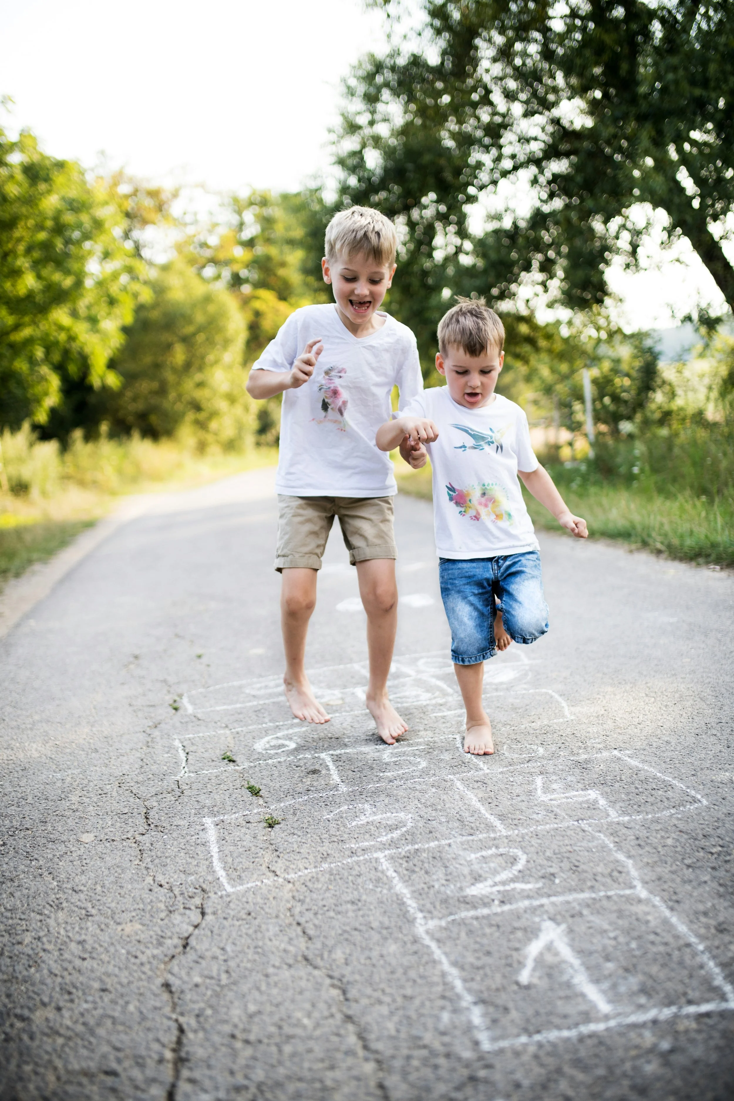 Two young boys playing hopscotch outdoors on a sunny day, surrounded by green trees.