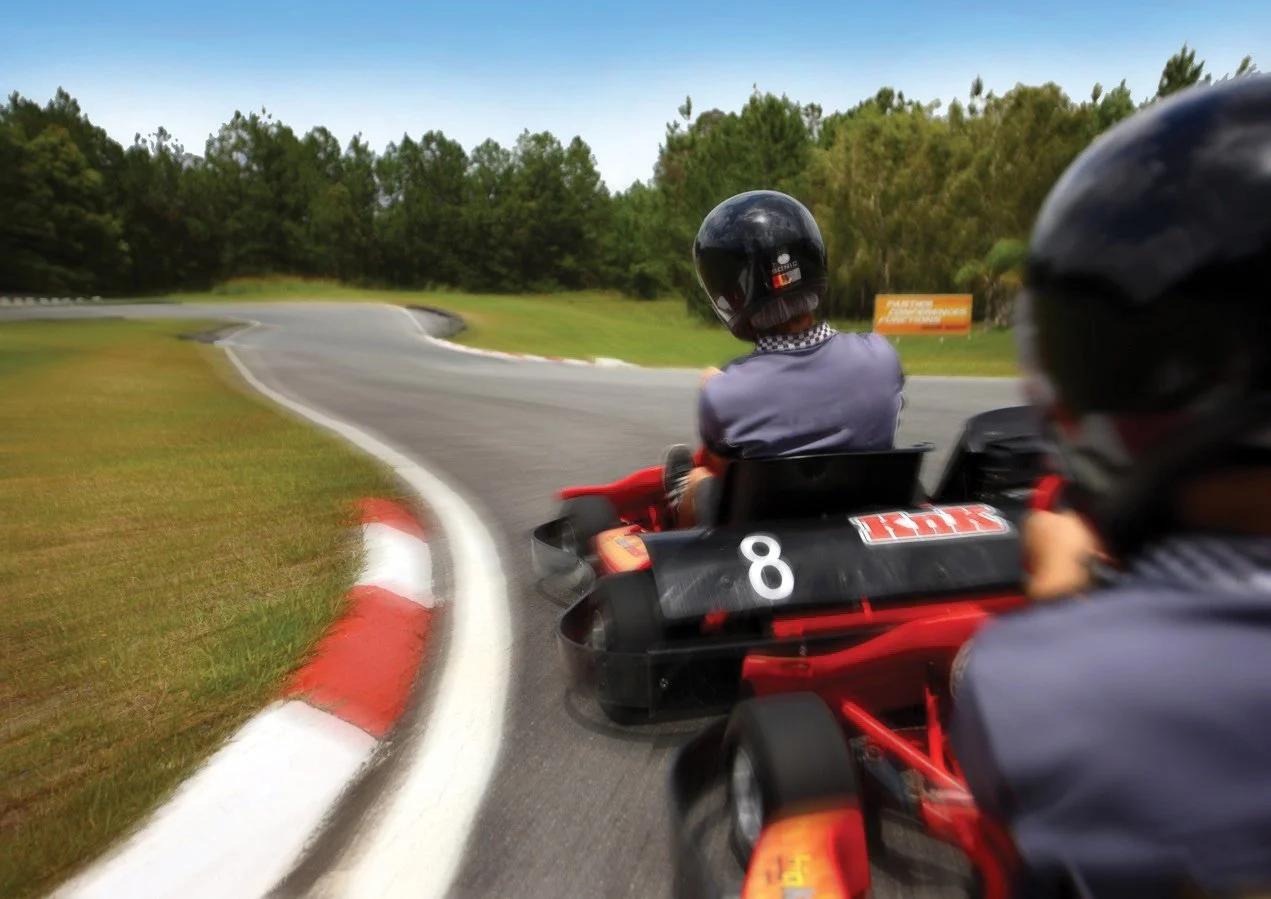 Two people in go-karts with helmets racing on a curving outdoor track surrounded by trees.