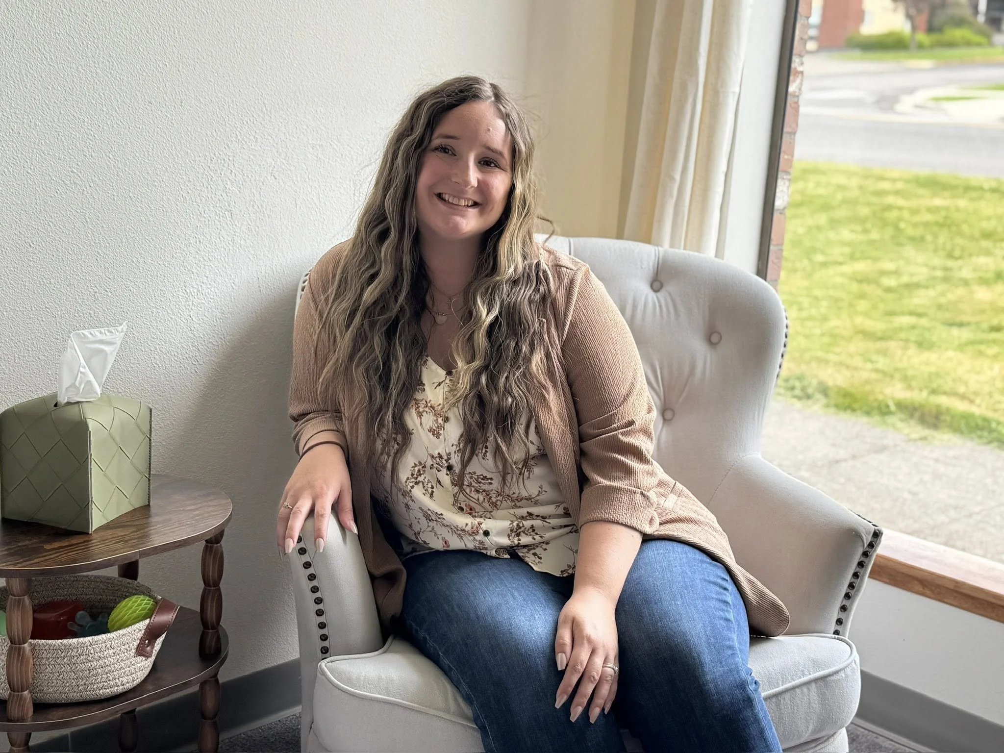 A woman with long wavy hair and a bright smile sitting on a white armchair next to a round wooden side table inside a home, with a window showing a lawn outside.