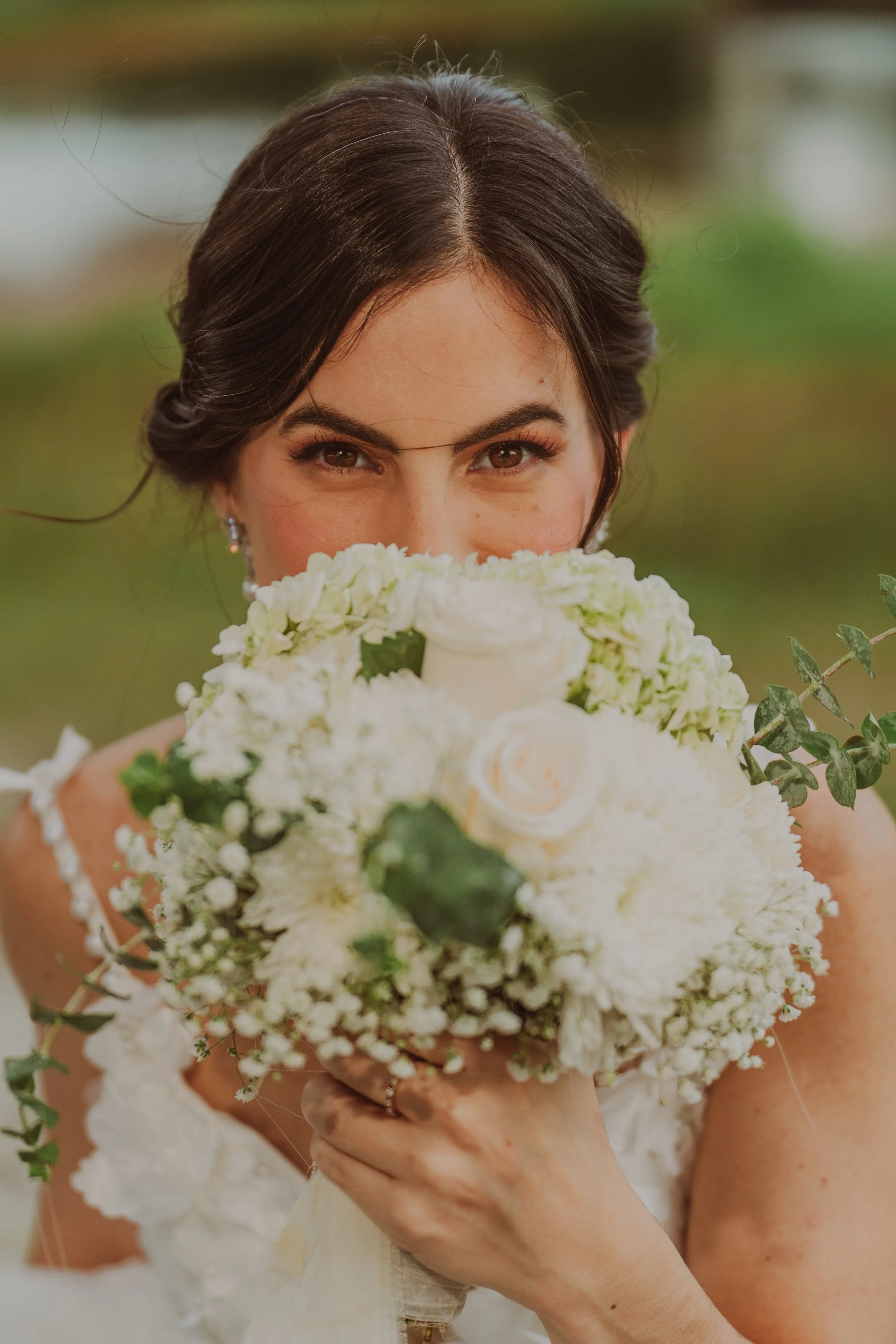 A bride with dark hair holding a large white floral bouquet, hiding her lower face and smiling with her eyes.