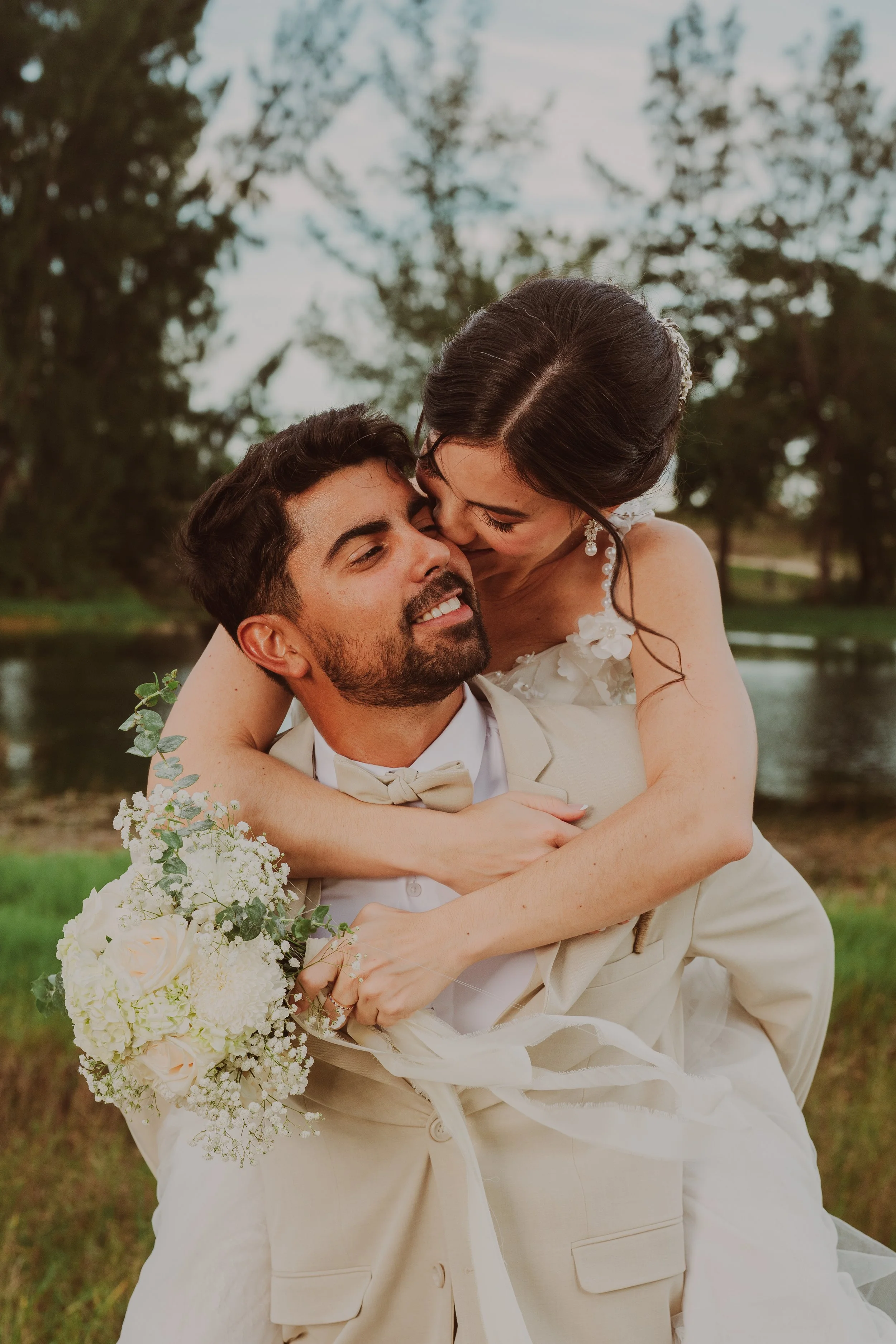 A bride and groom in wedding attire, outdoors near a river, sharing a joyful moment with the bride embracing the groom and holding a bouquet of white flowers.