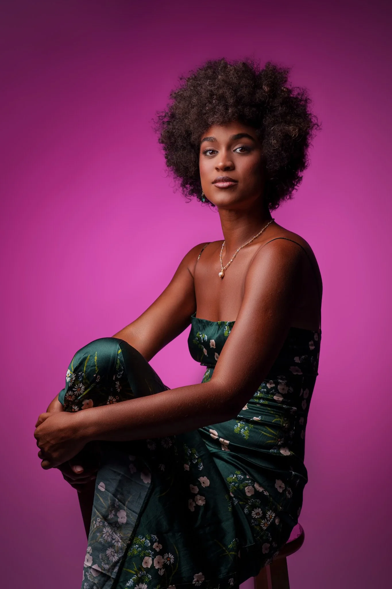 A woman with dark skin and curly hair sitting on a stool against a pink gradient background. She is wearing a floral dress and gold jewelry, looking confidently at the camera.