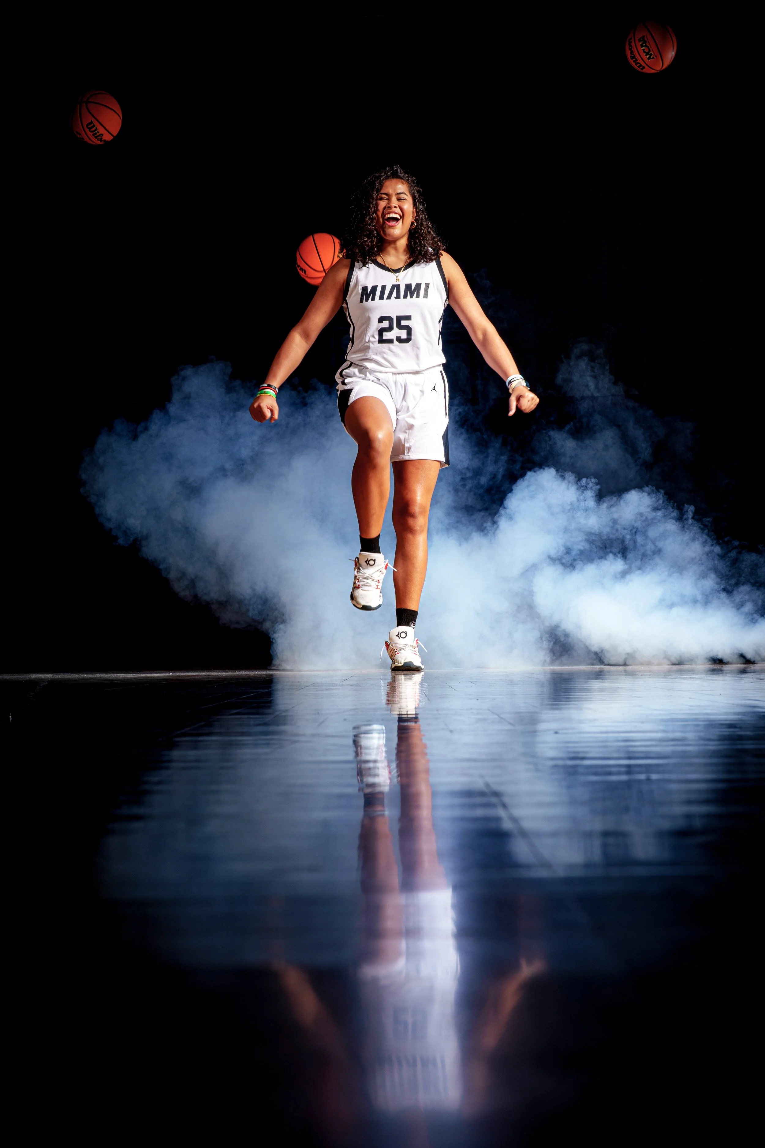 Female basketball player wearing a Miami jersey with the number 25 is walking confidently on a reflective floor with smoke and basketballs in the background.