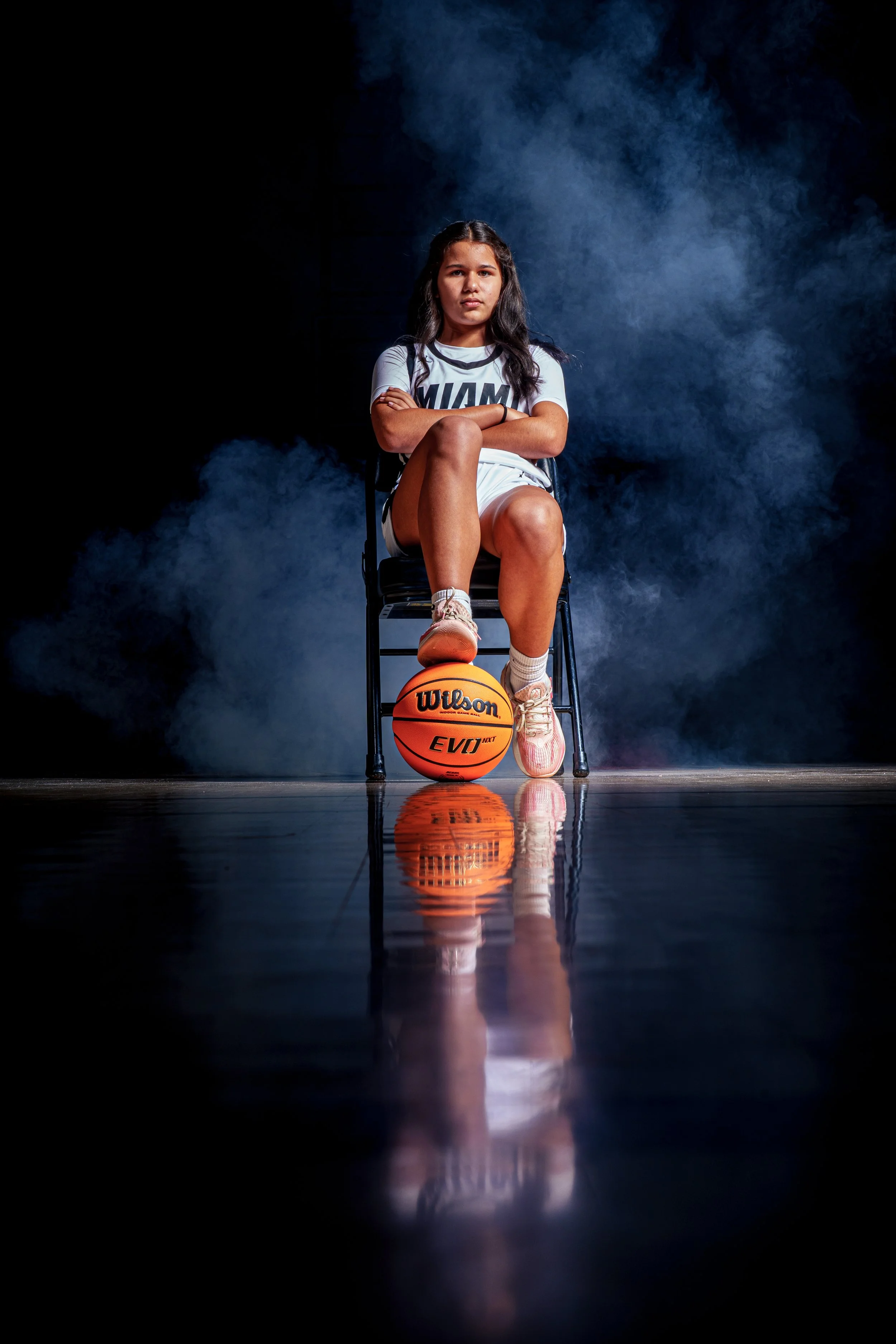 A female basketball player sitting on a chair with crossed arms, wearing a uniform with 'MIAMI' on it, her left foot resting on a Wilson basketball, on a reflective gym floor with dark background and smoke.