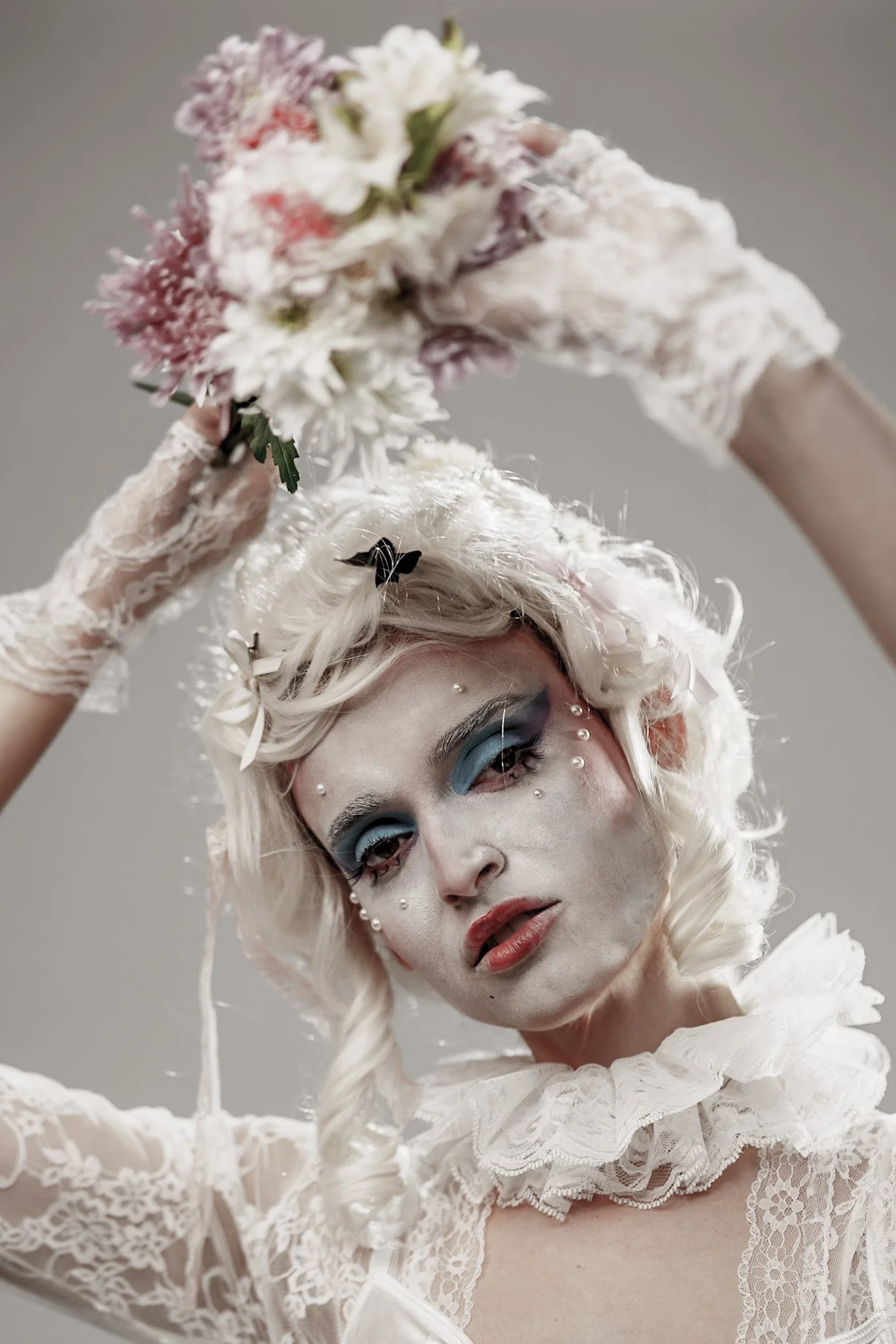 A person with platinum blonde curly hair, wearing a lacy white top and gloves, holding a bouquet of flowers above their head, with dramatic makeup including blue eyeshadow and rhinestones around their eyes.