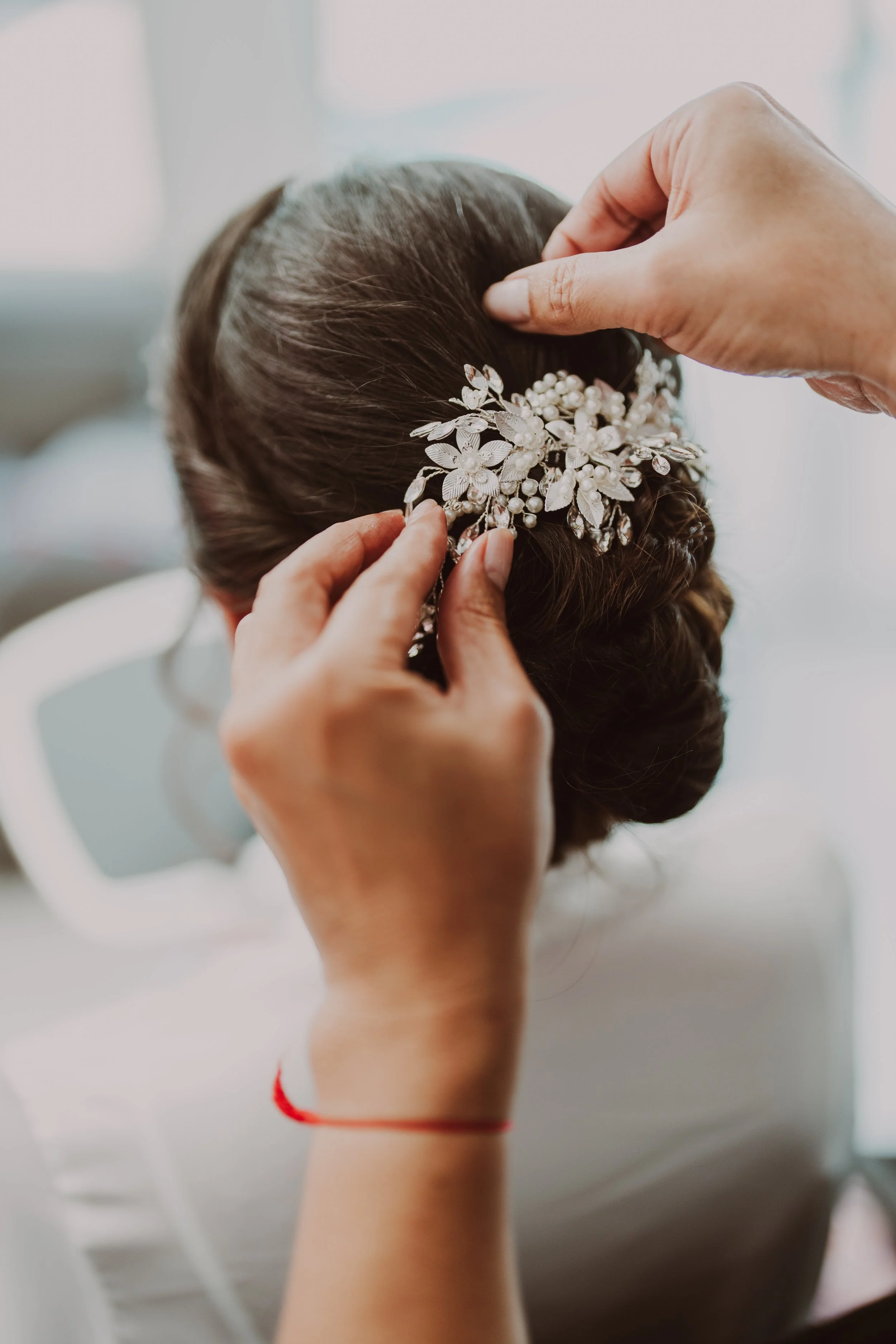 A woman with dark hair is getting her hair adorned with a decorative floral hairpiece with pearls, as another person styles her hair.