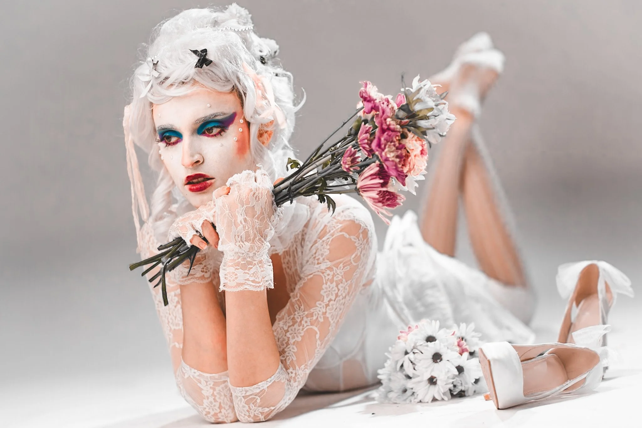 A woman with radical makeup and curly platinum blonde hair lying on her stomach on a light gray background, holding pink and white flowers, dressed in a lace outfit and high heels.