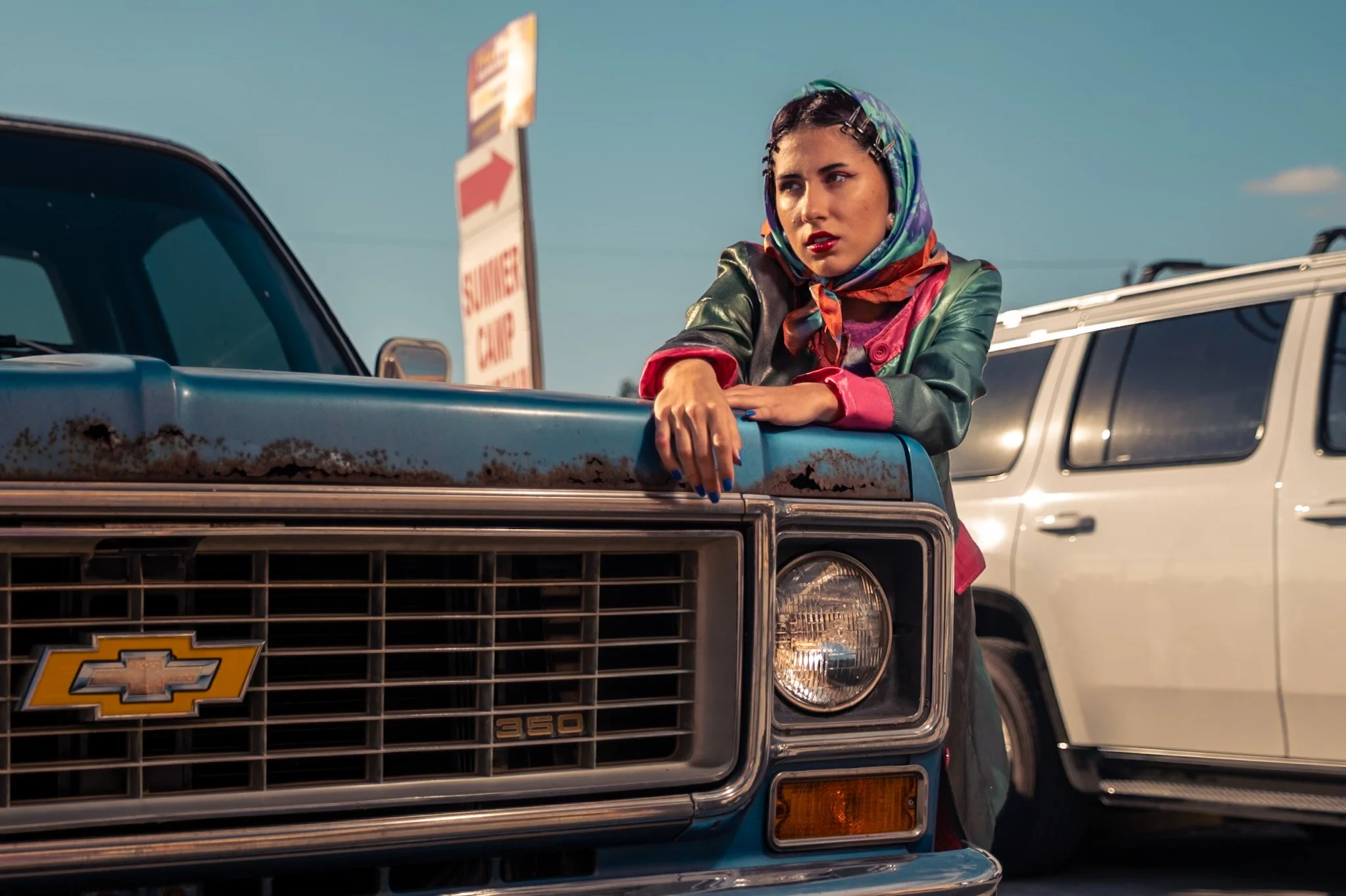 A woman leaning on the hood of a blue Chevrolet truck, with a white vehicle behind her, under a clear blue sky.