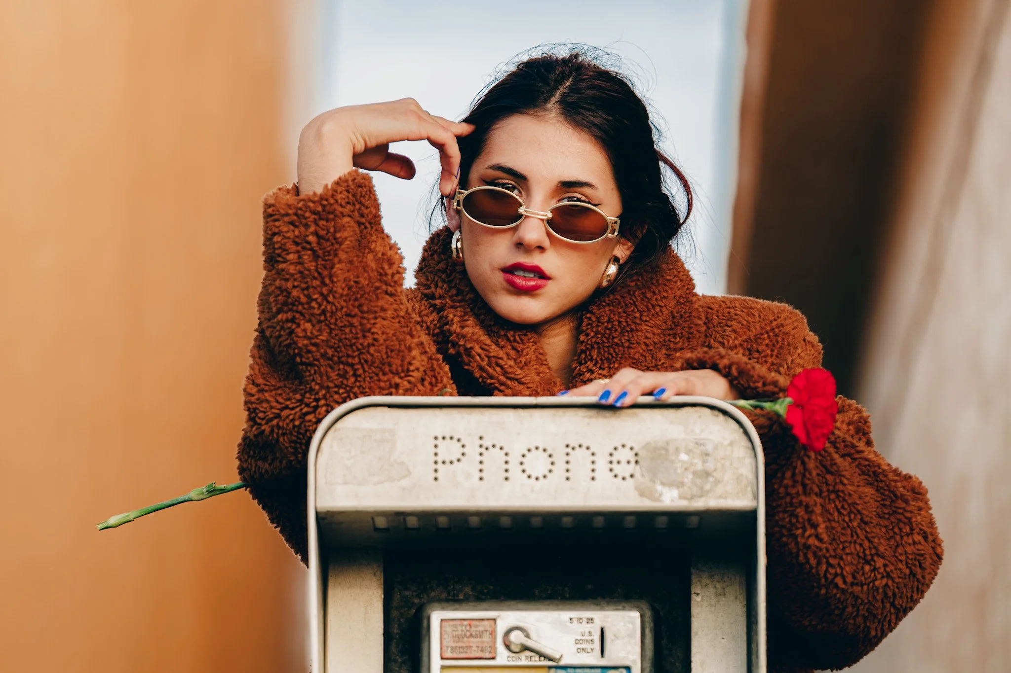 A woman with dark hair and wearing sunglasses, a brown fuzzy coat, and earrings, is leaning on an old, dusty payphone with a red flower in her hand, looking at the camera.