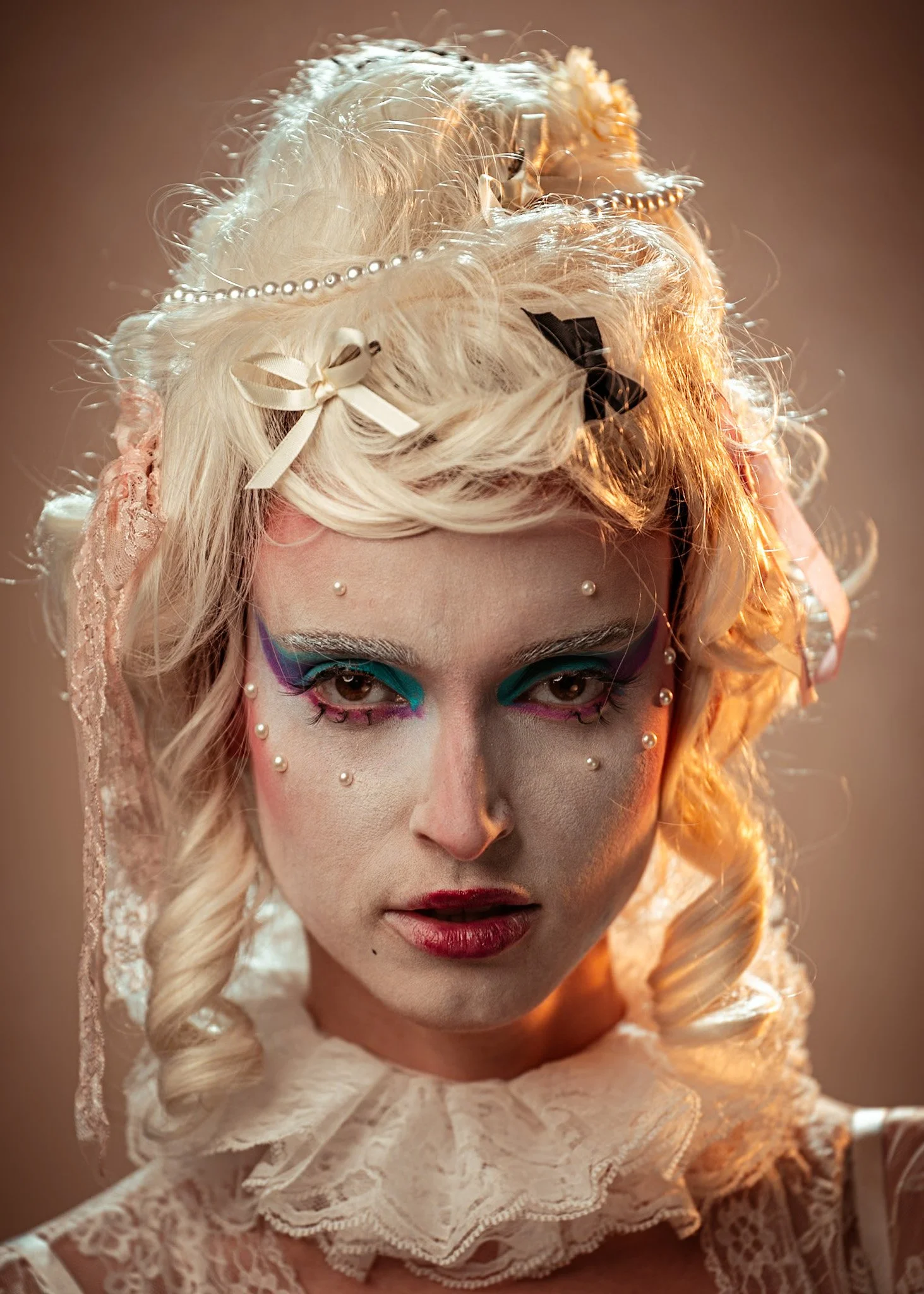 A woman with elaborate makeup, blonde curled hair adorned with ribbons, pearls, and lace, wearing a lace choker, and with various pearls and makeup accents on her face.