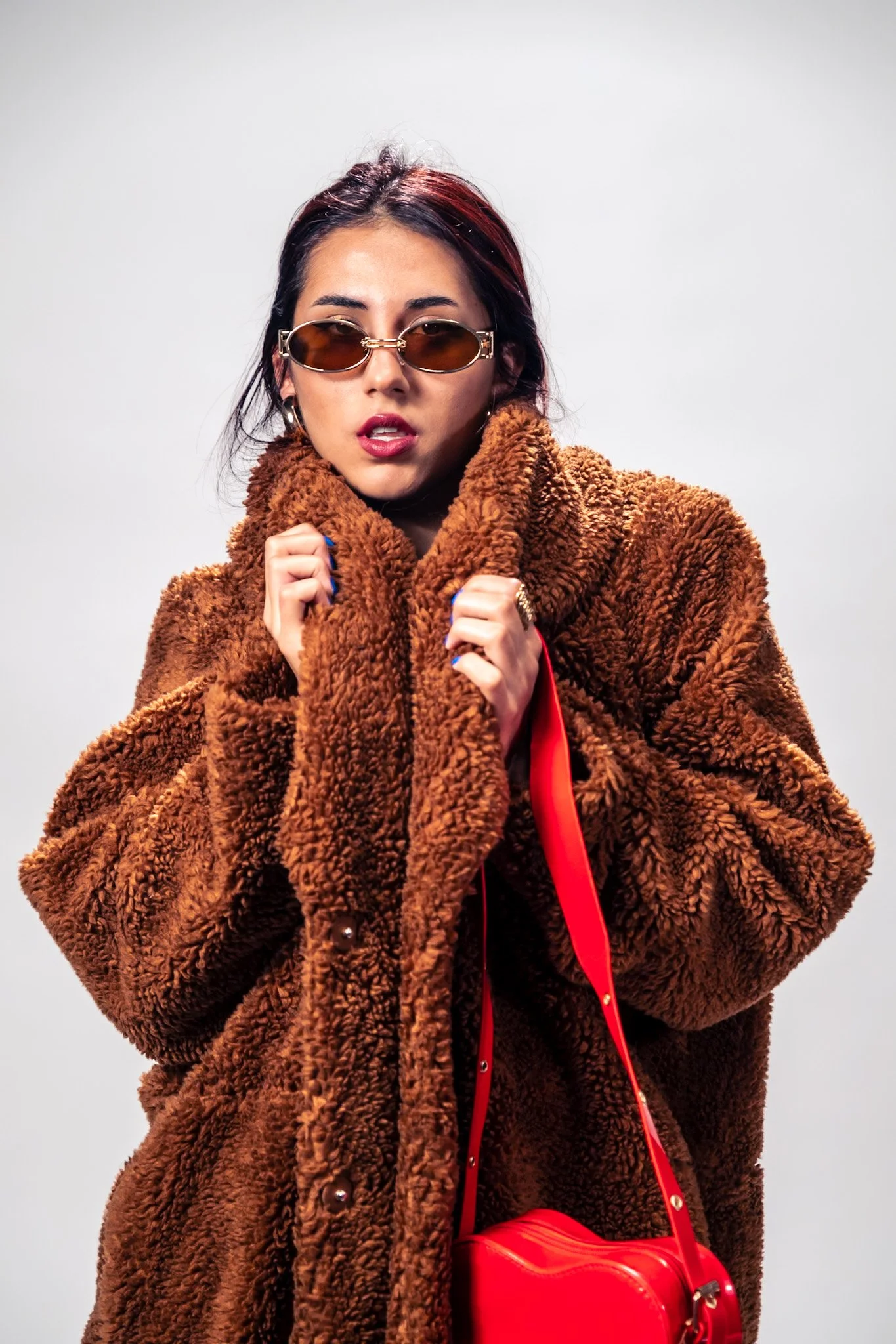 A woman wearing sunglasses, a large brown fuzzy coat, and holding a red purse against a plain gray background.