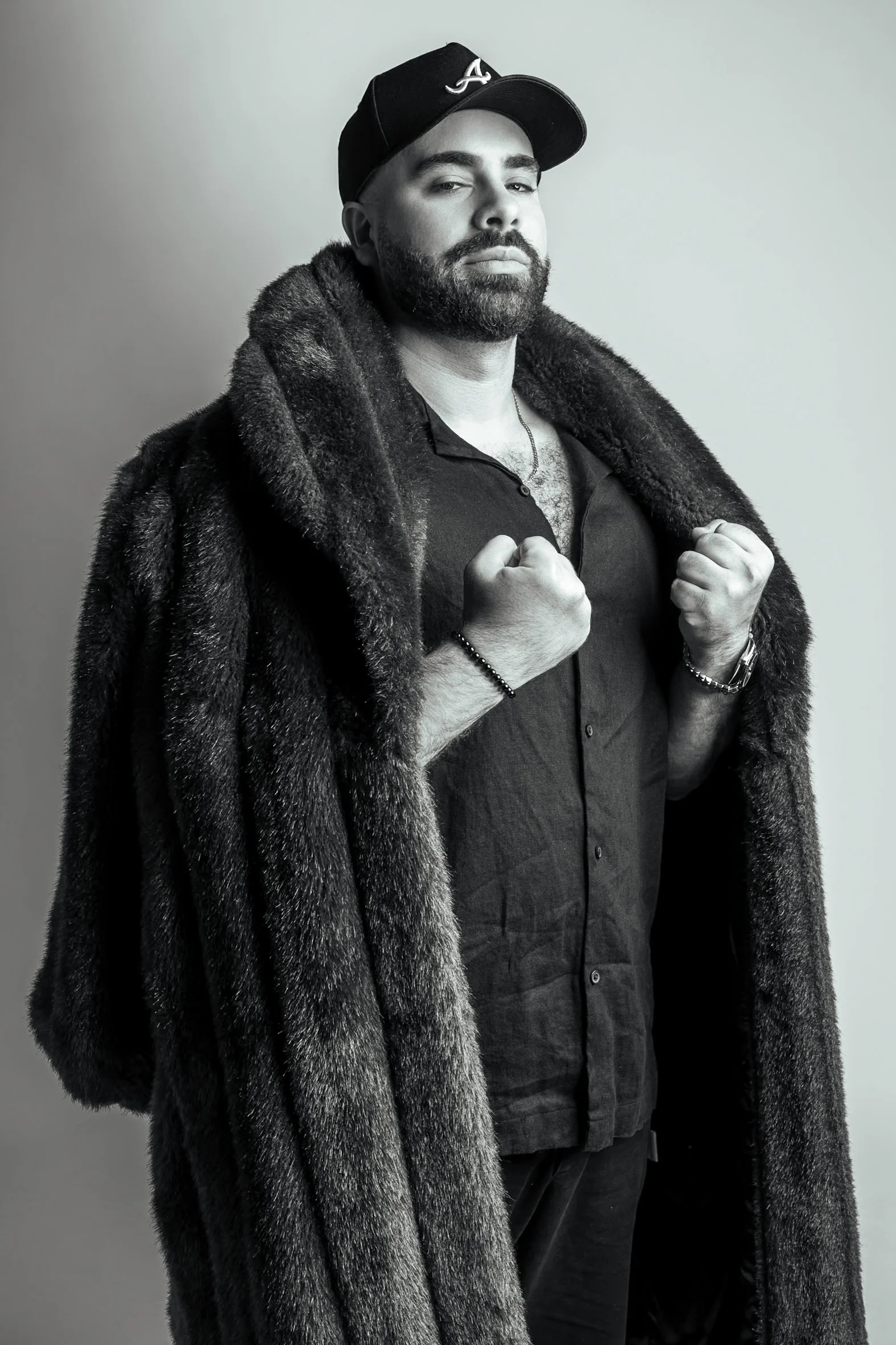A man with a beard wearing a black baseball cap with an 'A' on it, a dark shirt, and a large fur coat. He is holding the lapels of his coat with fists raised slightly, looking confidently at the camera.