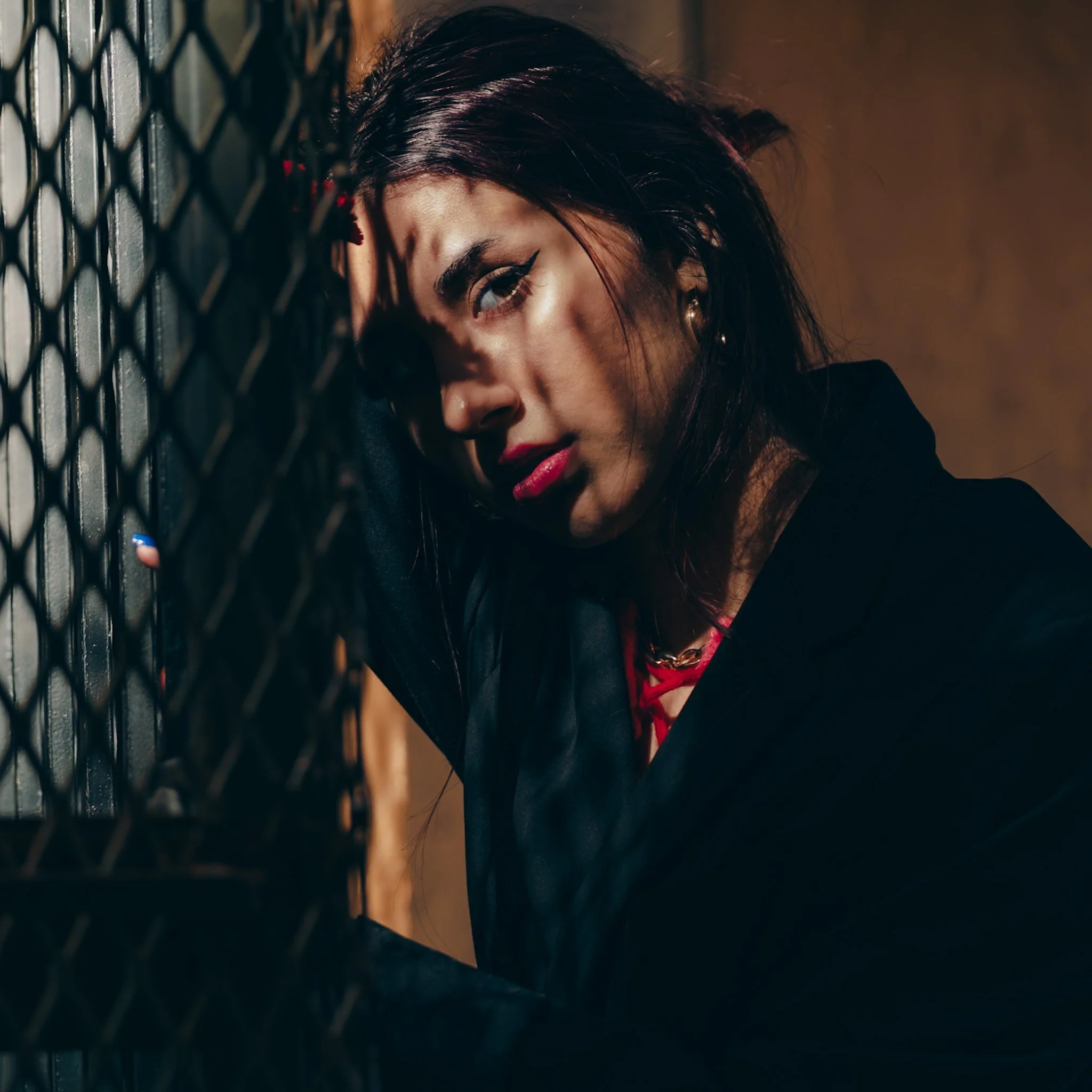 A woman with dark hair and bold makeup leaning against a metal grid with an intense expression.