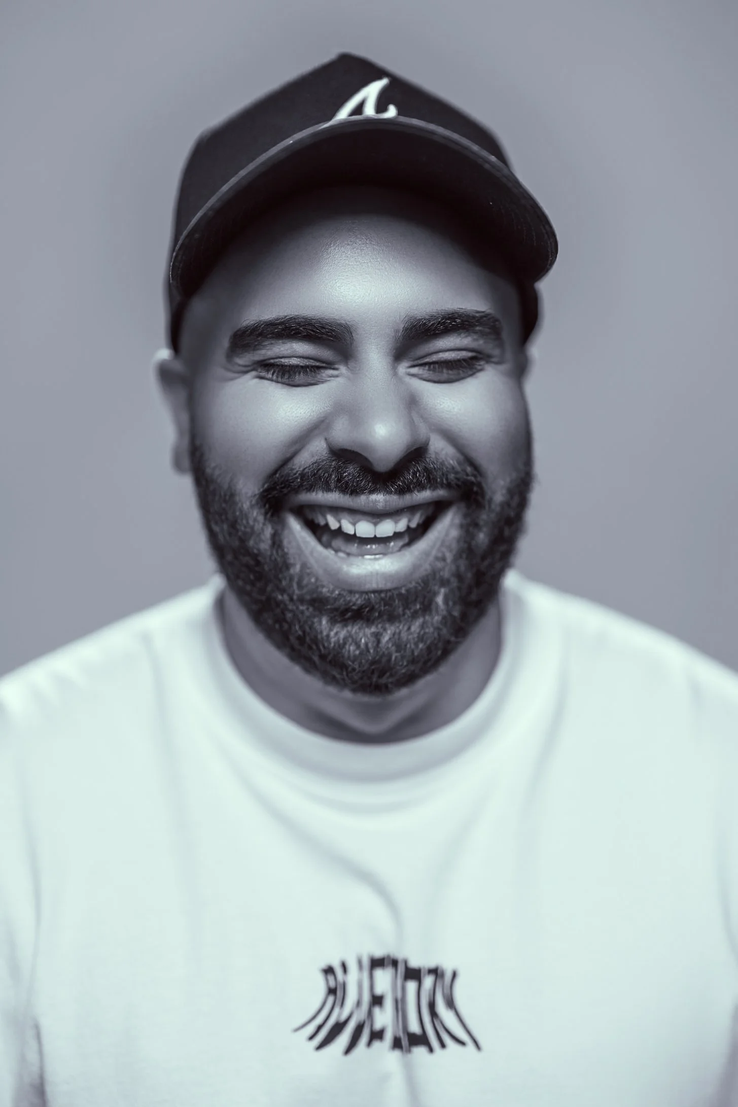 A black and white photo of a smiling man with a beard wearing a baseball cap and a white t-shirt.