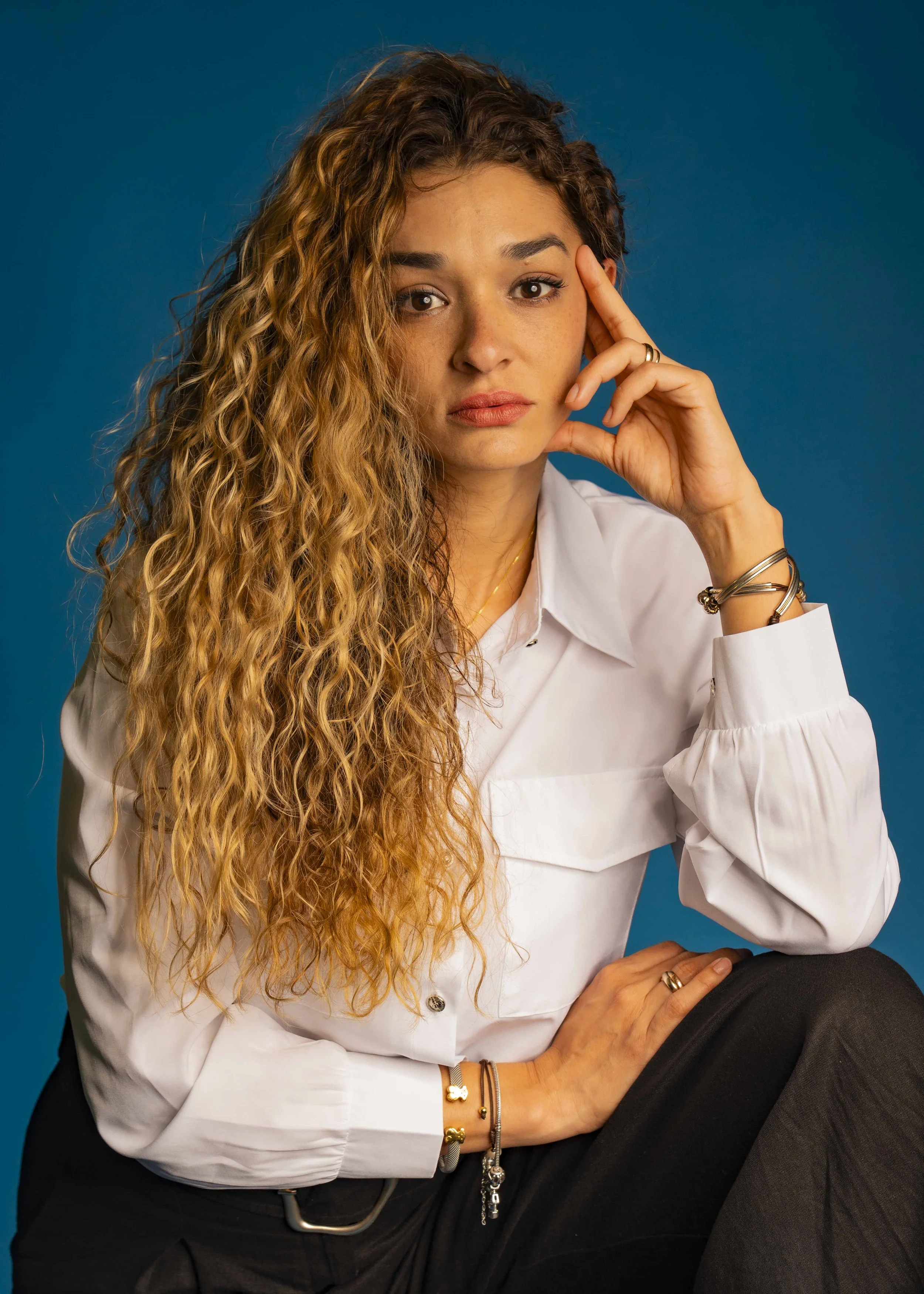 A woman with long, curly hair wearing a white shirt and black pants, sitting against a blue background, looking serious with her hand near her temple.
