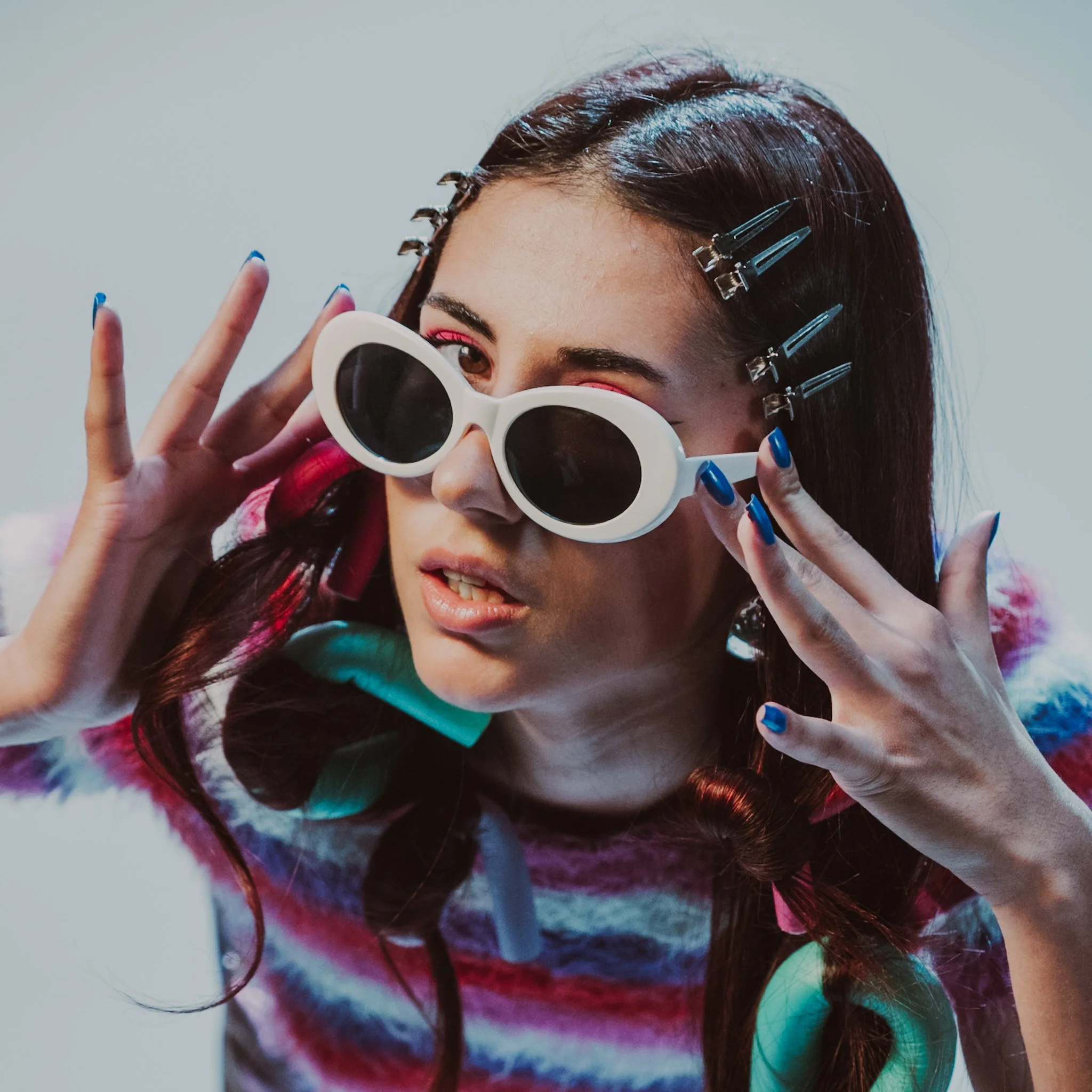 A young woman with dark hair, wearing large white sunglasses, colorful striped shirt, and holding her hands near her face with nails painted blue. She has hair clips in her hair and a playful expression.