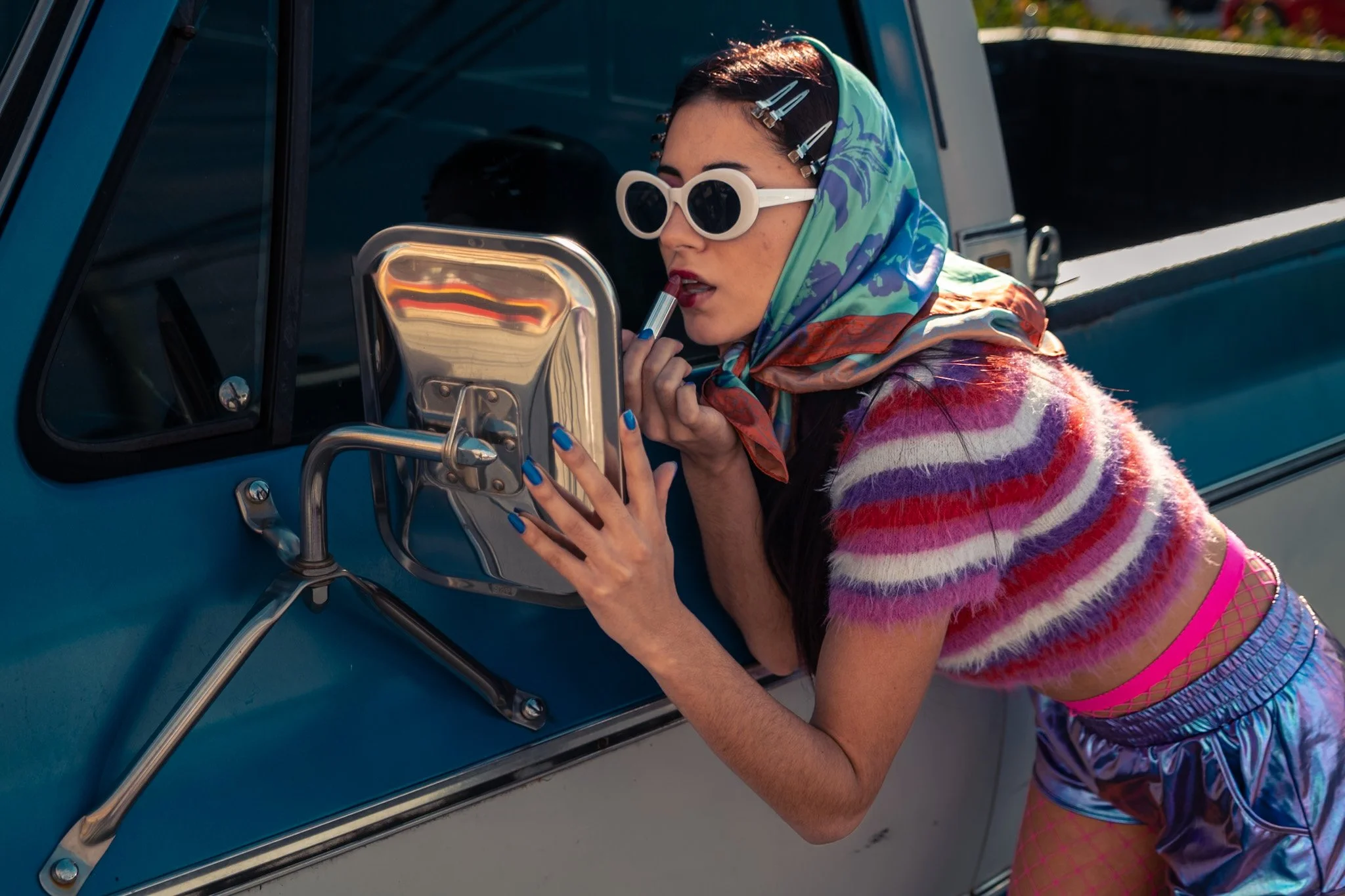 A woman with dark hair, wearing large white sunglasses, colorful scarf, striped furry top, pink fishnet shorts, and shiny blue pants, applies lipstick while looking in a mirror attached to a vintage blue truck.