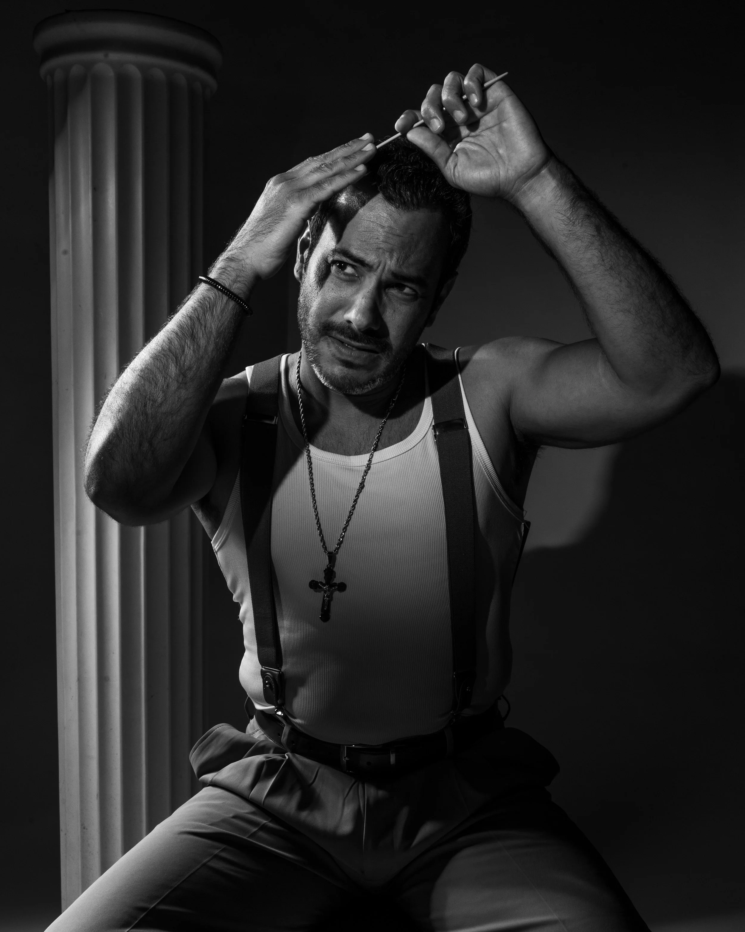 A man with a serious facial expression holding an acupuncture needle in his forehead, sitting next to a classical column, wearing a tank top with suspenders and a cross necklace, in black and white.