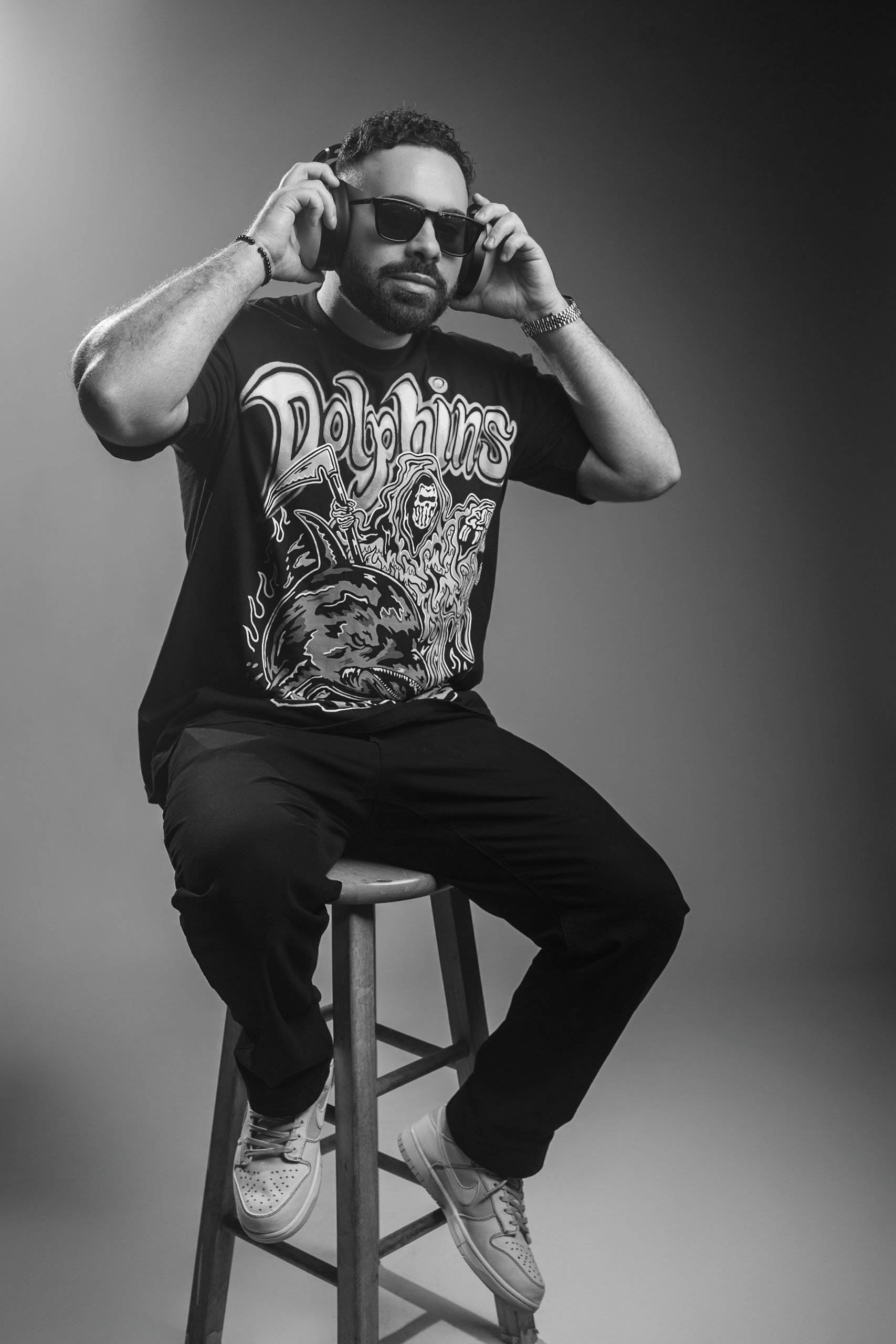 A man sitting on a wooden stool, wearing sunglasses, headphones, a graphic T-shirt, black pants, and sneakers, in a studio with a plain backdrop.