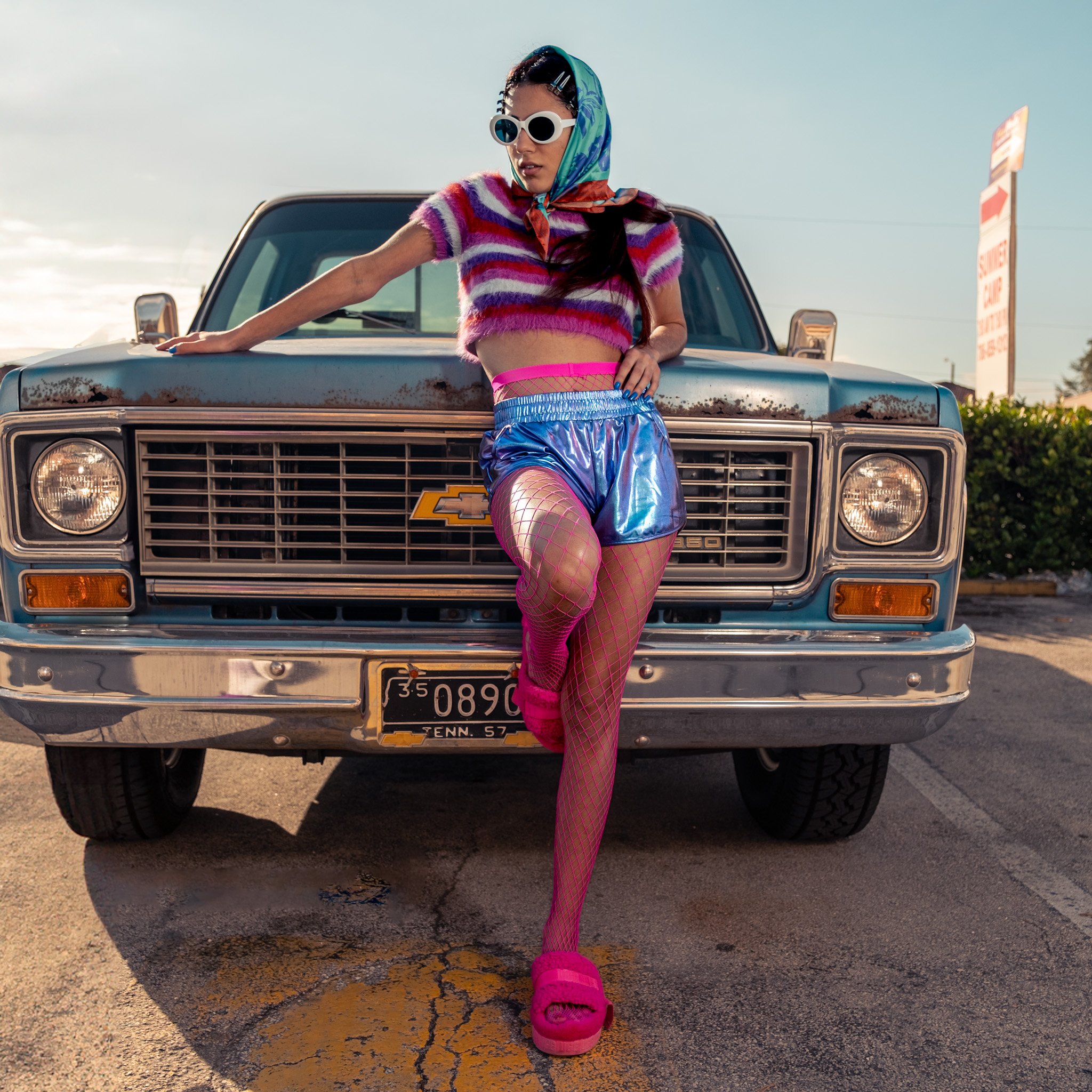 A woman in colorful, retro-inspired clothing leaning against the hood of a vintage blue pickup truck in a parking lot. She is wearing a rainbow striped crop top, shiny blue shorts, bright pink fishnet stockings, pink platform shoes, and large white s