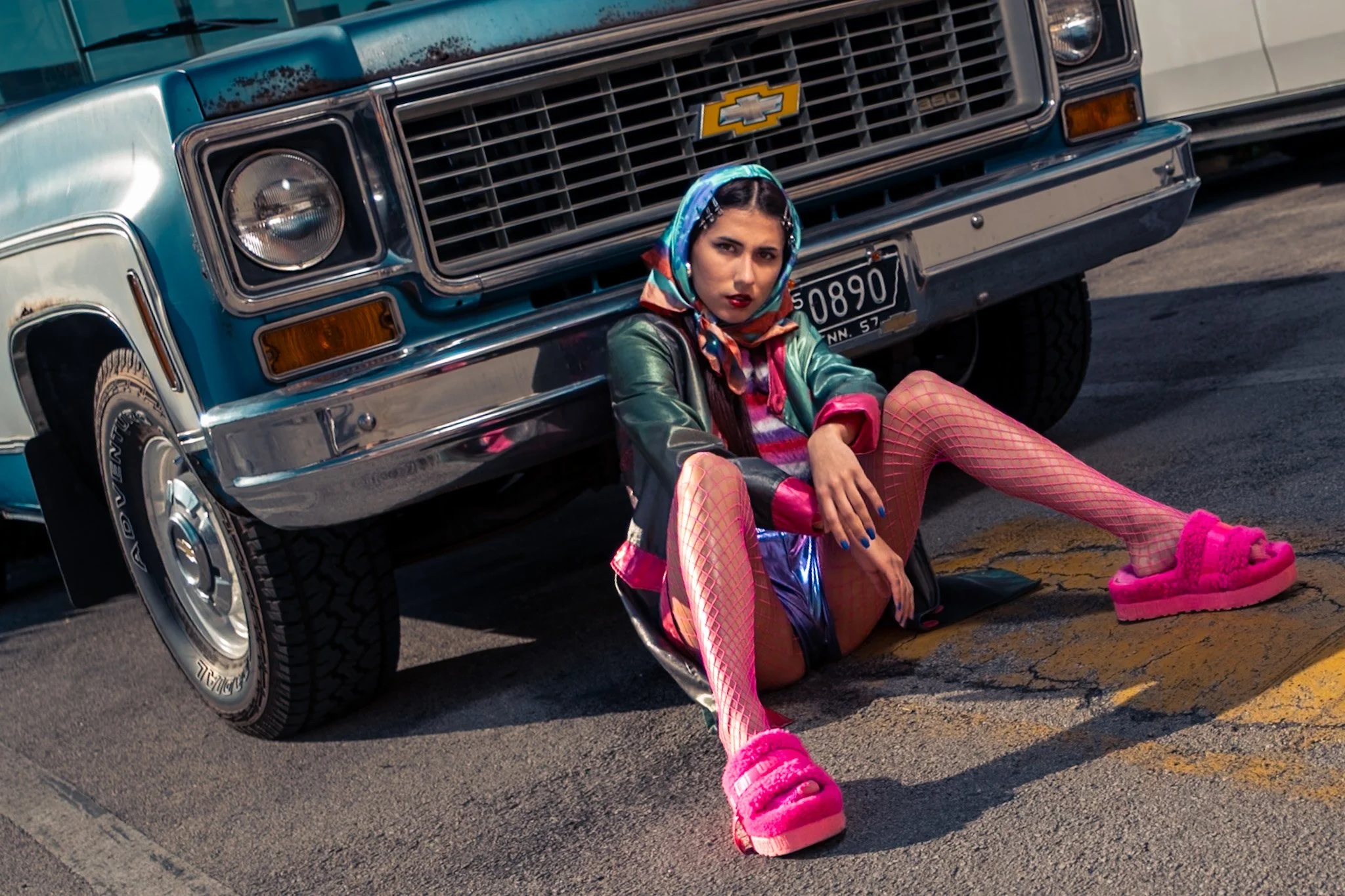 A woman sitting on the ground in front of a blue vintage Chevrolet truck, wearing colorful clothing, fishnet stockings, bright pink slippers, a patterned headscarf, and dark lipstick.