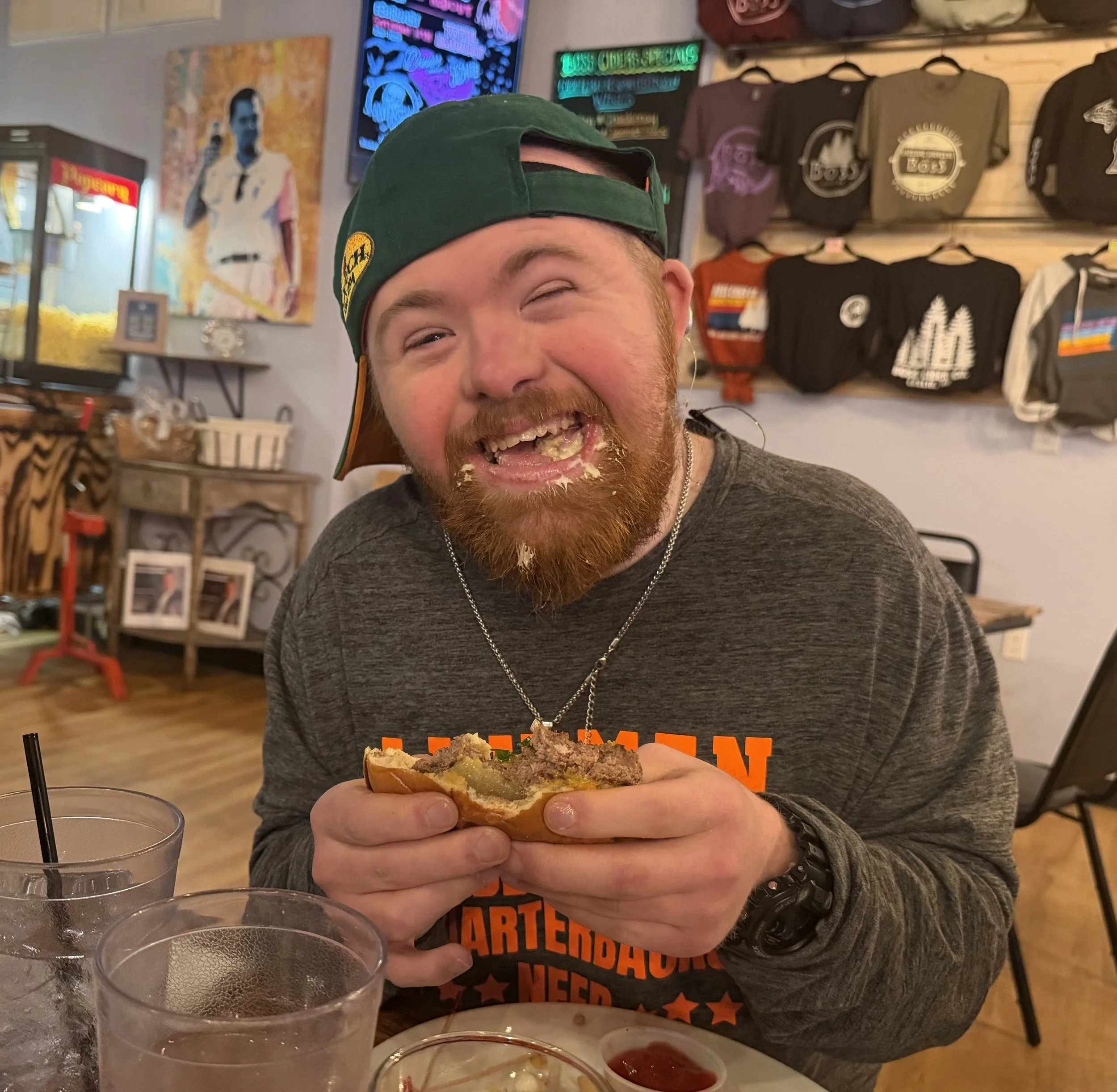 A man with a red beard and mustache wearing a green baseball cap backwards, eating a burger with messy toppings, smiling broadly with food on his face, sitting in a restaurant or cafe.