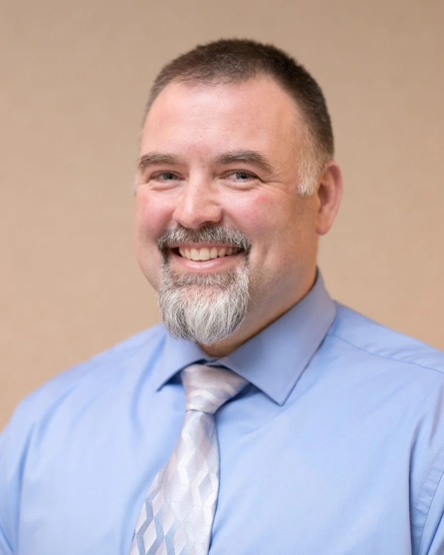 A smiling middle-aged man with short, dark hair, a beard with gray, wearing a light blue dress shirt and a patterned tie, against a plain beige background.