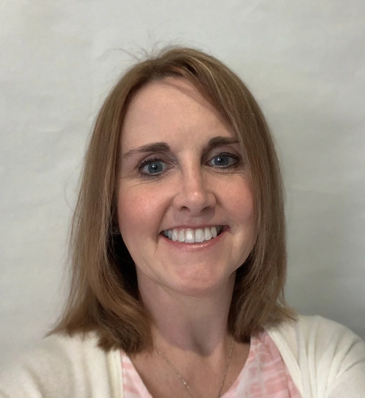 Close-up of a smiling woman with shoulder-length reddish-brown hair, blue eyes, wearing a light-colored top, against a plain white wall.