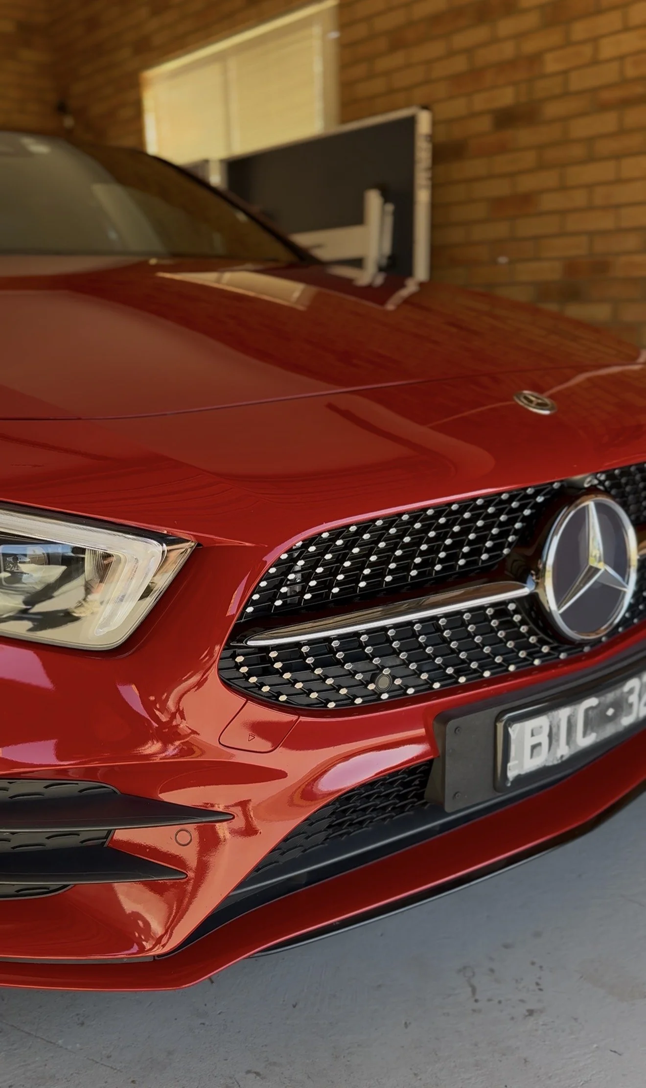 Close-up of a red Mercedes-Benz car, showing the front grille, headlight, and part of the hood, with a brick wall and window in the background.