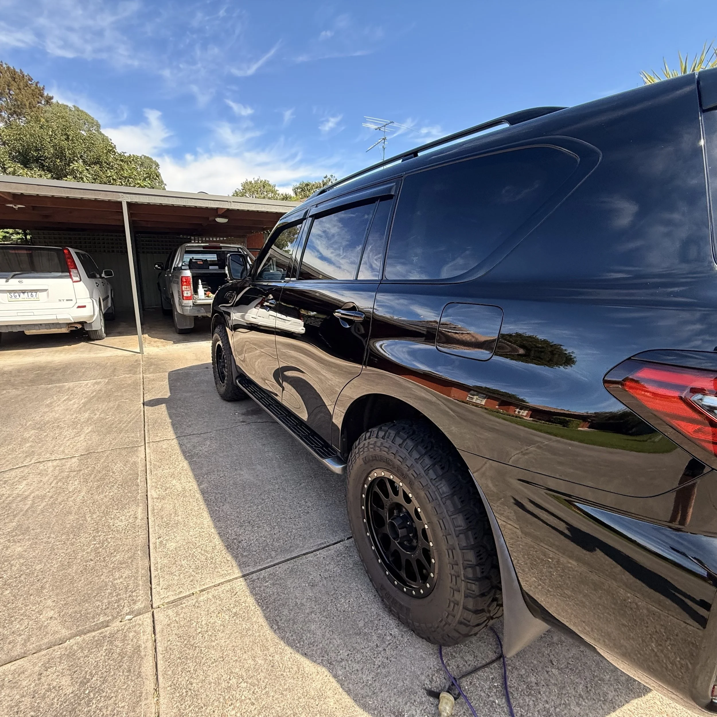 Black SUV parked in a driveway with a carport. Several other vehicles are parked nearby. The sky is blue with some clouds. The area is bordered by trees.