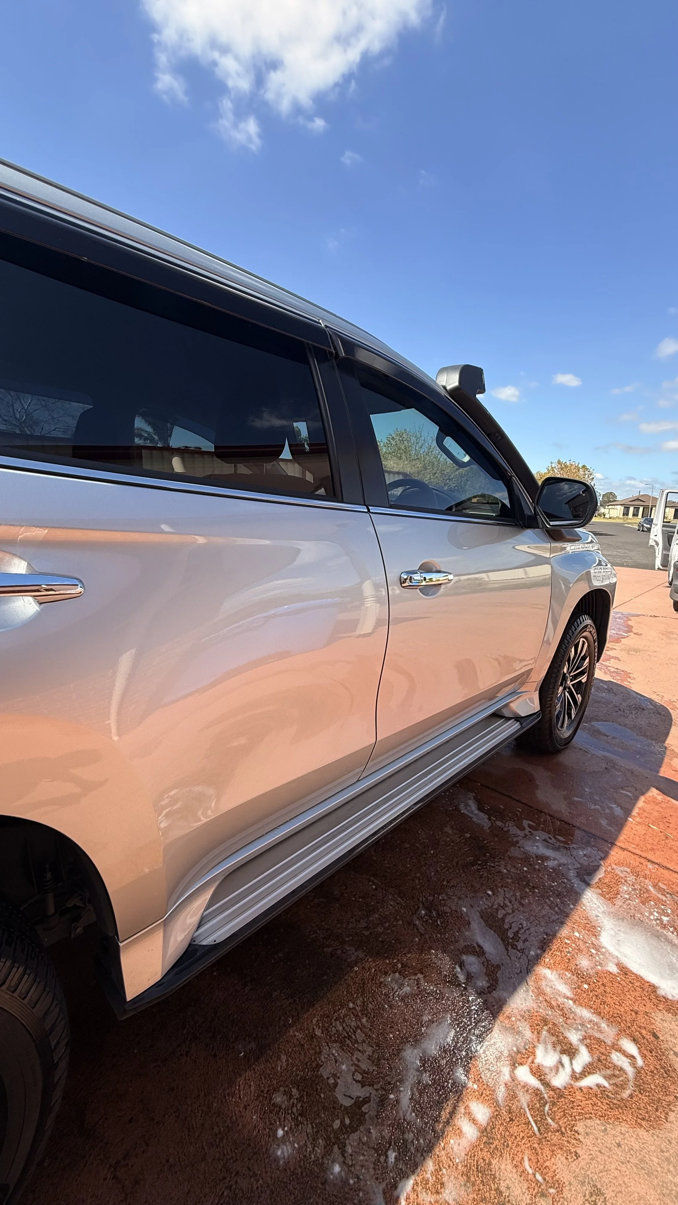 A silver SUV parked on a wet, red concrete surface outdoors with soap suds on the ground, under a blue sky with some clouds and a distant building.