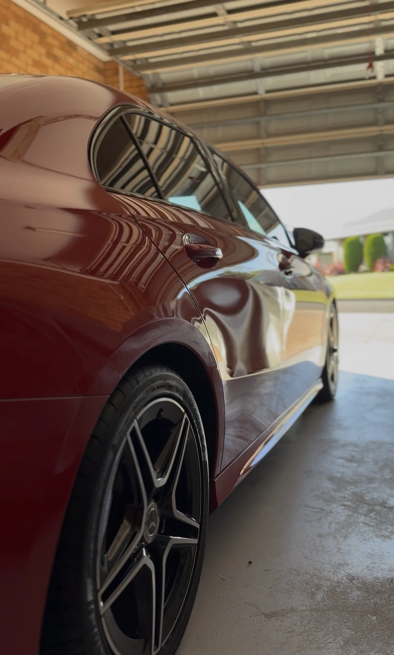 A close-up view of a shiny burgundy sports car parked in a garage, showing the rear side and part of the front, with reflections of the garage's ceiling and ceiling beams visible on its surface.