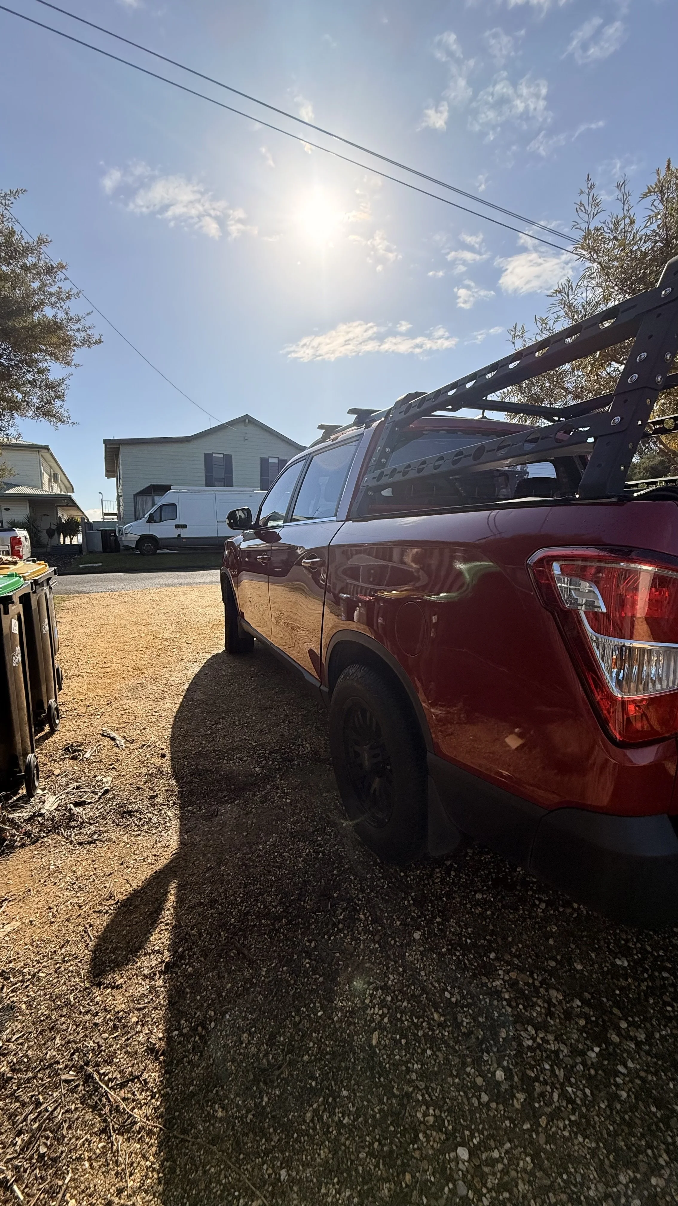 A red pickup truck parked on a gravel driveway under a sunny sky with some clouds. In the background, there are houses and a white van parked across the street.