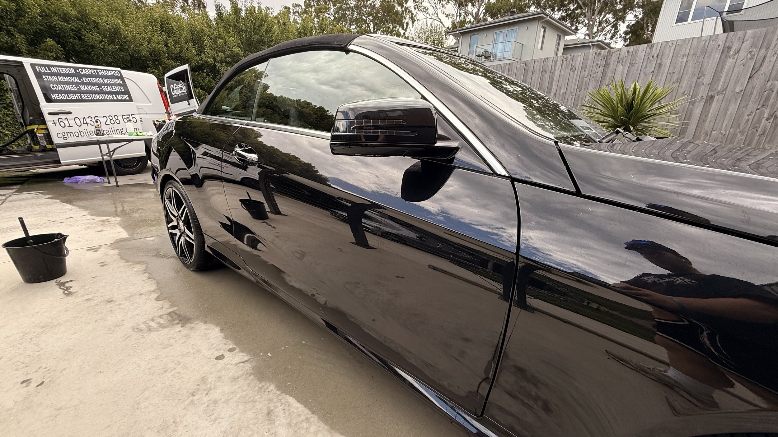 A black car is parked in a driveway with cleaning supplies nearby, including a bucket and a sprayer. In the background, there is a white van with advertising for detailing services, and a residential area with a house and a wooden fence.