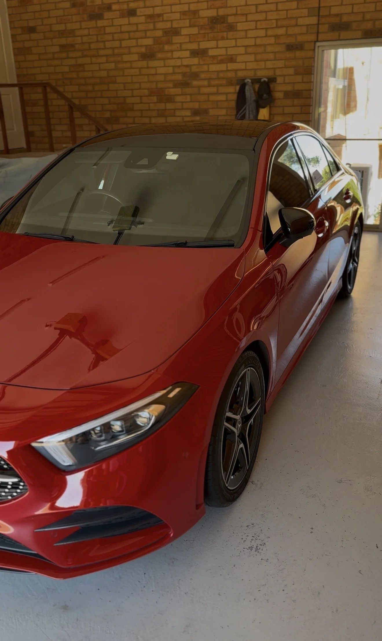 Red sports car parked inside a garage with brick walls and a window with sunlight coming through. There are two jackets hanging on the brick wall in the background.
