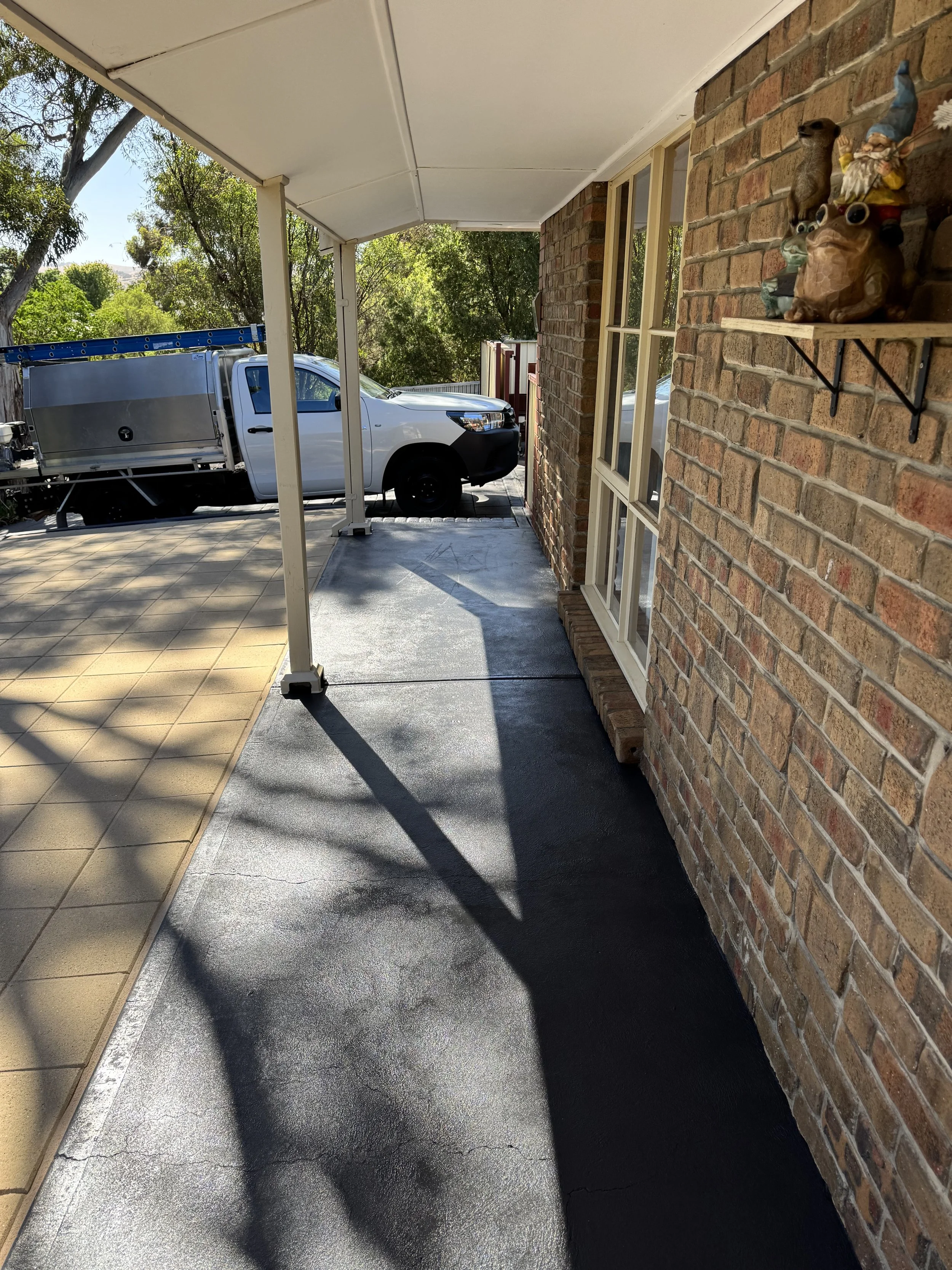 A covered porch area with a brick exterior wall decorated with ceramic figurines and shadowed from nearby trees, a driveway with a parked white utility truck, and a background of green trees and blue sky.