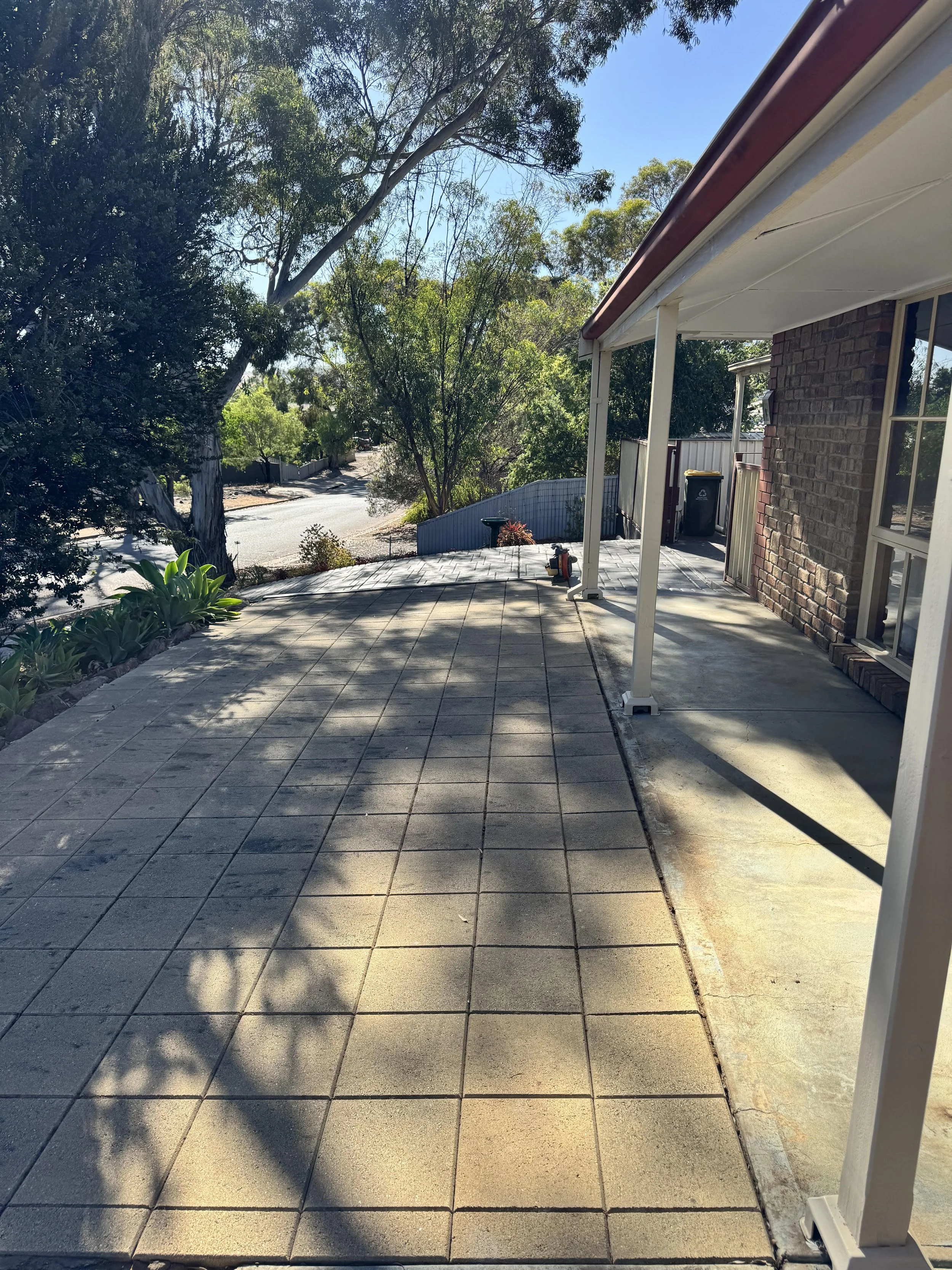 Front porch of a house with patterned concrete tiles, a brick wall, and a white support beam, with trees and a street visible in the background under a clear blue sky.