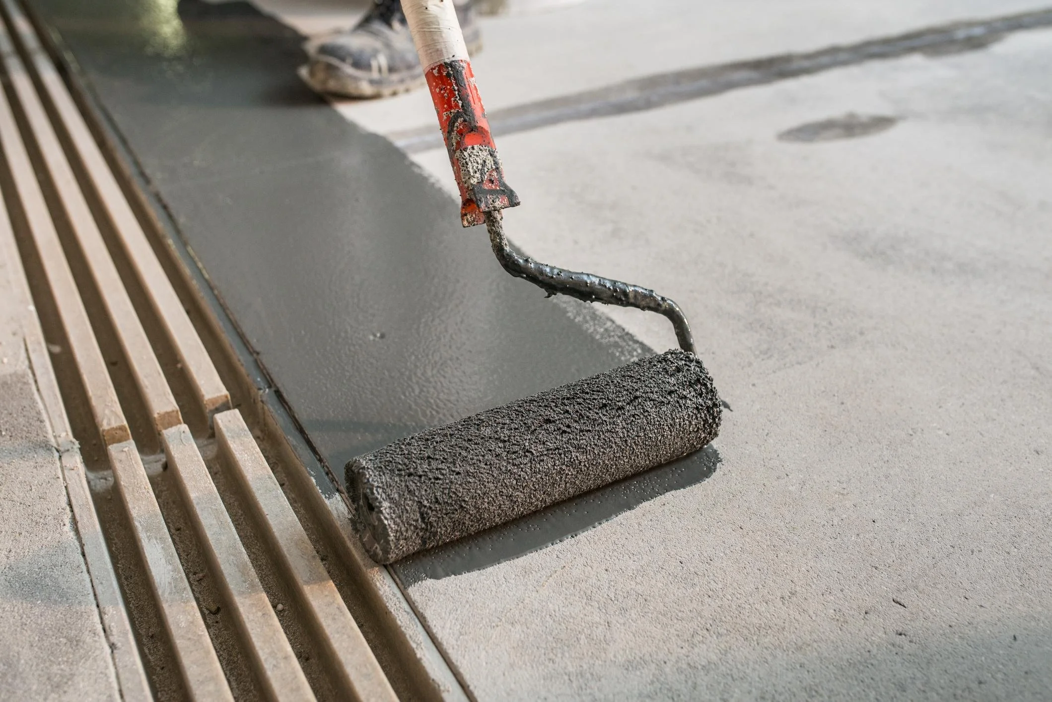A trowel with a red handle and black mud on it next to a sponge, spreading gray concrete on a surface, with a drain grate nearby.