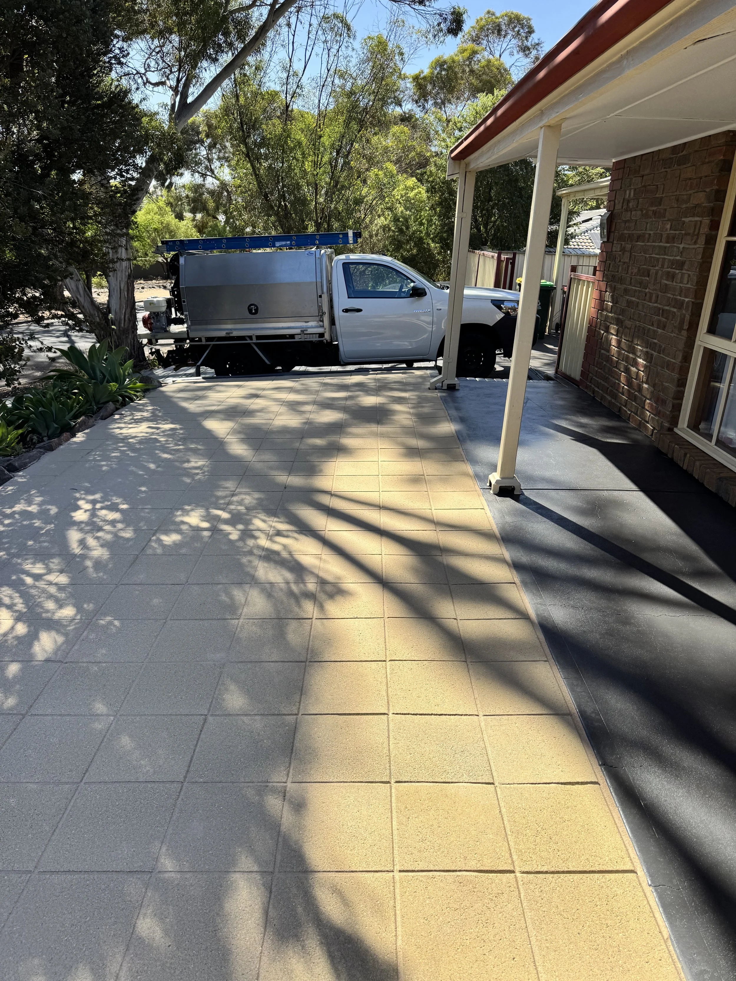A white utility truck parked on a concrete patio next to a brick house with trees in the background and a blue sky.