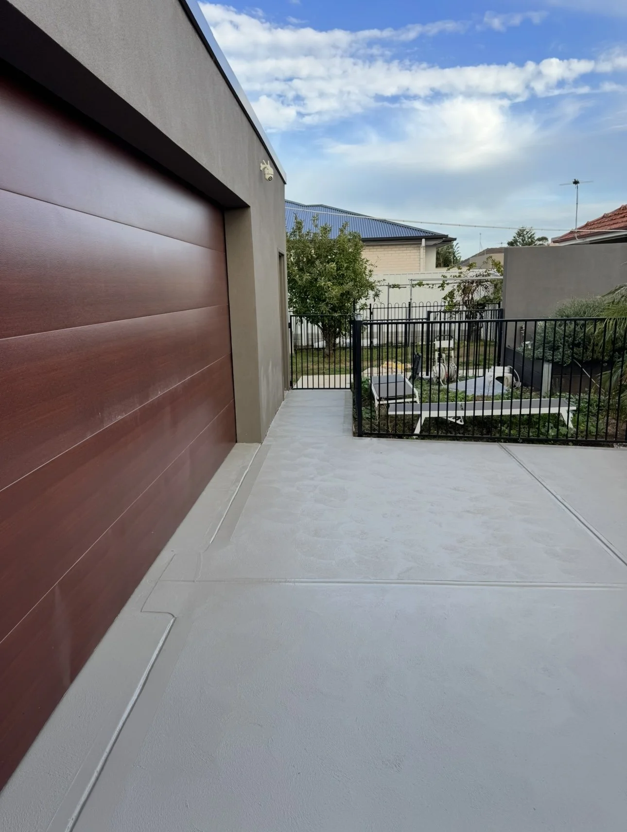 A backyard patio with a white concrete floor, black metal fence, and a view of trees, houses with blue and red roofs, and a partly cloudy sky.