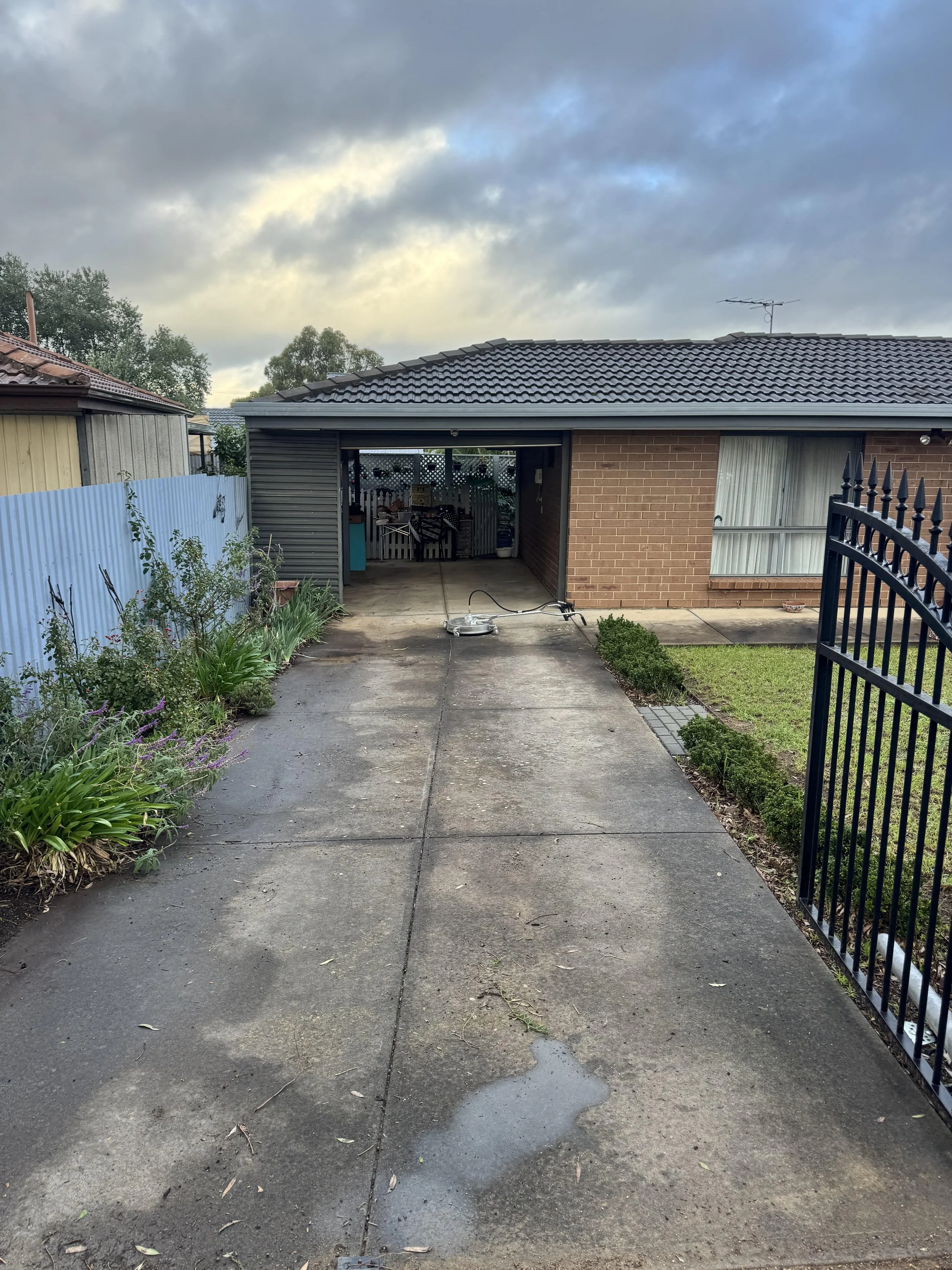 A backyard with a concrete driveway leading to a garage with an open entrance, brick and metal siding, and a gray tiled roof. There are plants and flowers on the left, a grass patch on the right, a black metal fence on the right side, and cloudy sky overhead.