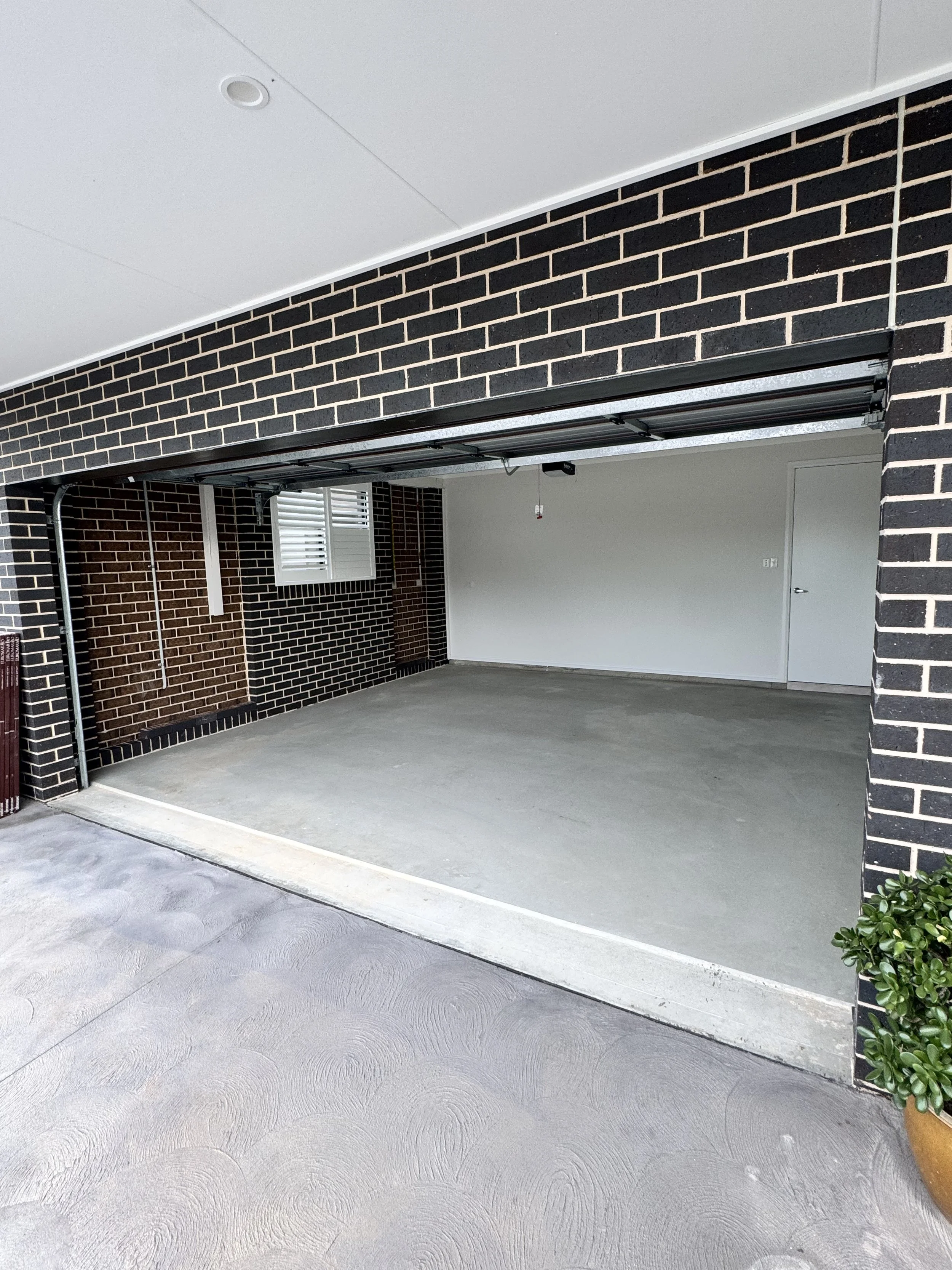 Empty garage with brick wall, white ceiling, concrete floor, window with shutters, and potted plant on the right.