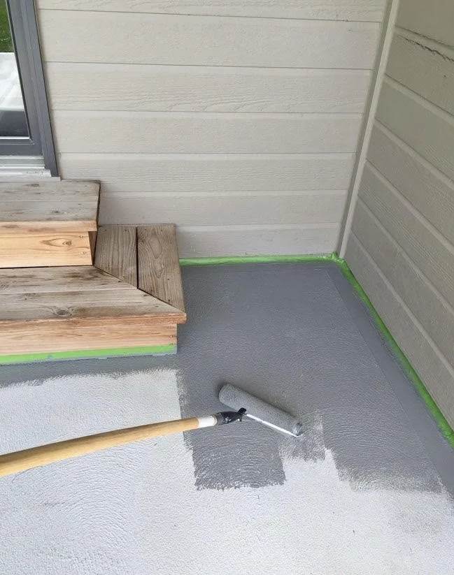 A door mat and two wooden steps outside a house, with a person applying gray sealant or paint to the concrete porch floor using a paint roller, with green painter's tape along the edges.
