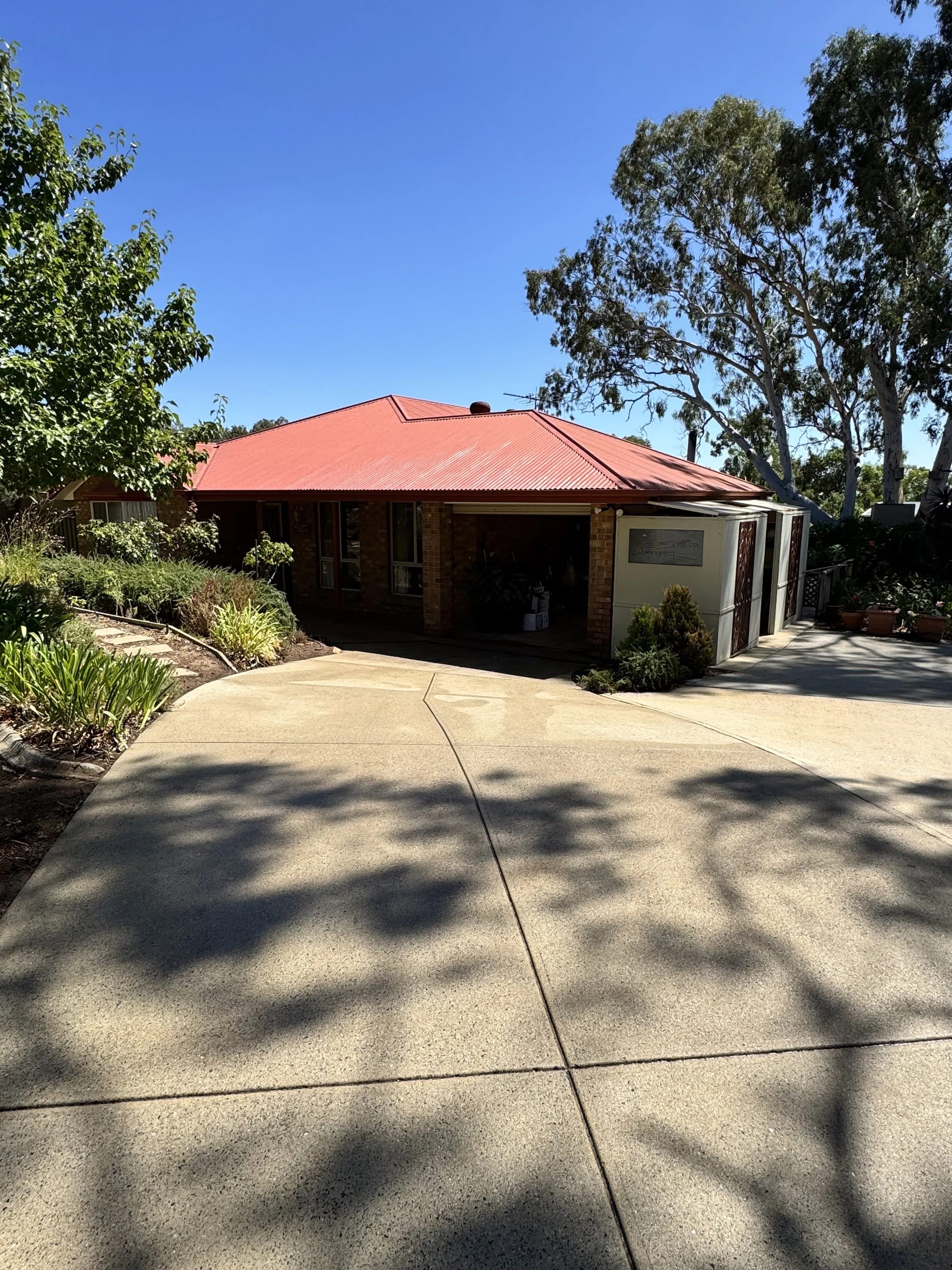 A house with a red metal roof surrounded by trees and plants, with a concrete driveway in the foreground and a clear blue sky overhead.