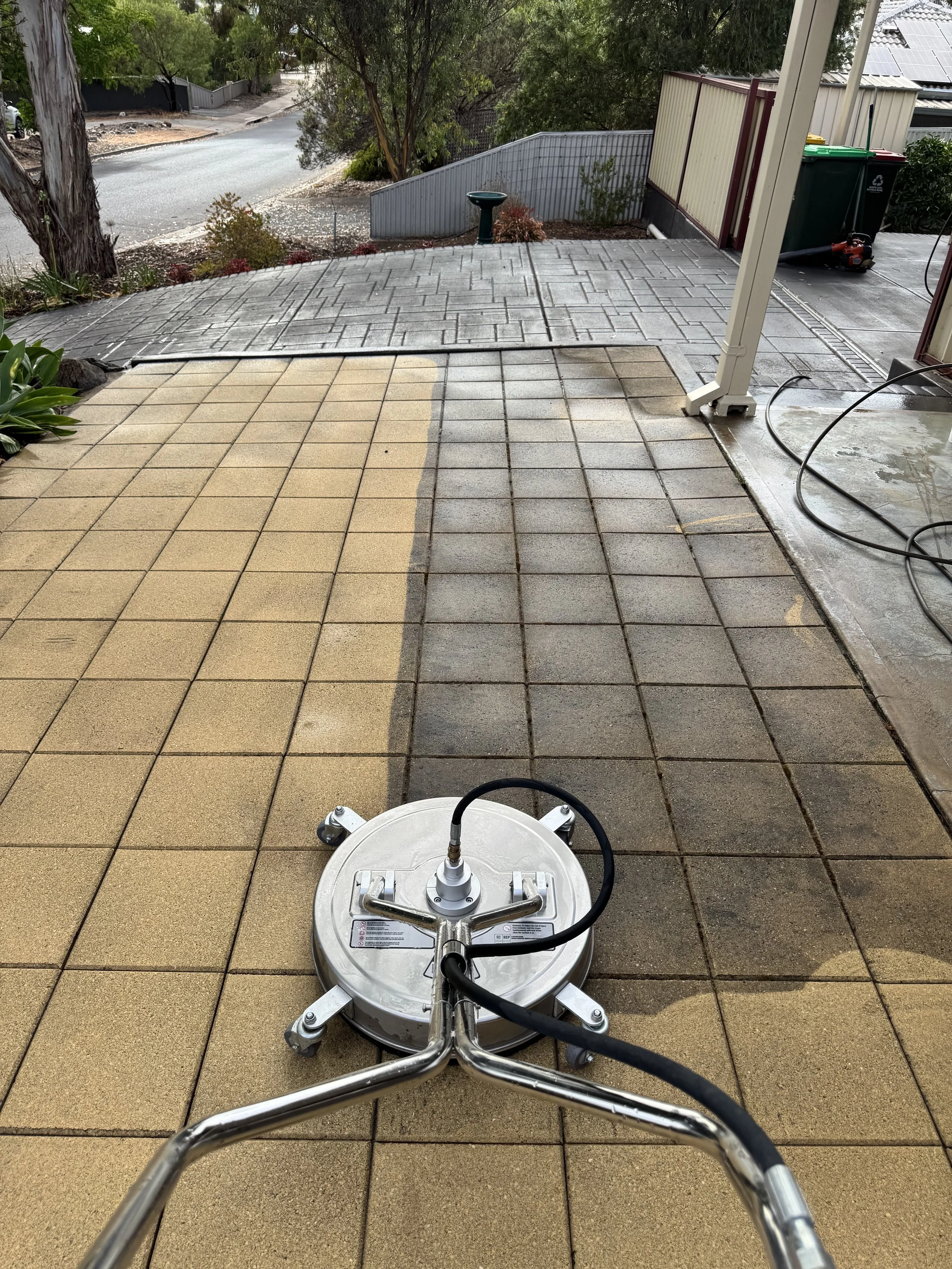 View from the front of a floor cleaning machine on a tiled patio with beige and gray tiles. The machine's metal handlebar and part of the cleaning head are visible. The background shows an outdoor area with a gray concrete paved area, a small water fountain, trees, shrubs, a road, and a house with solar panels on the roof.
