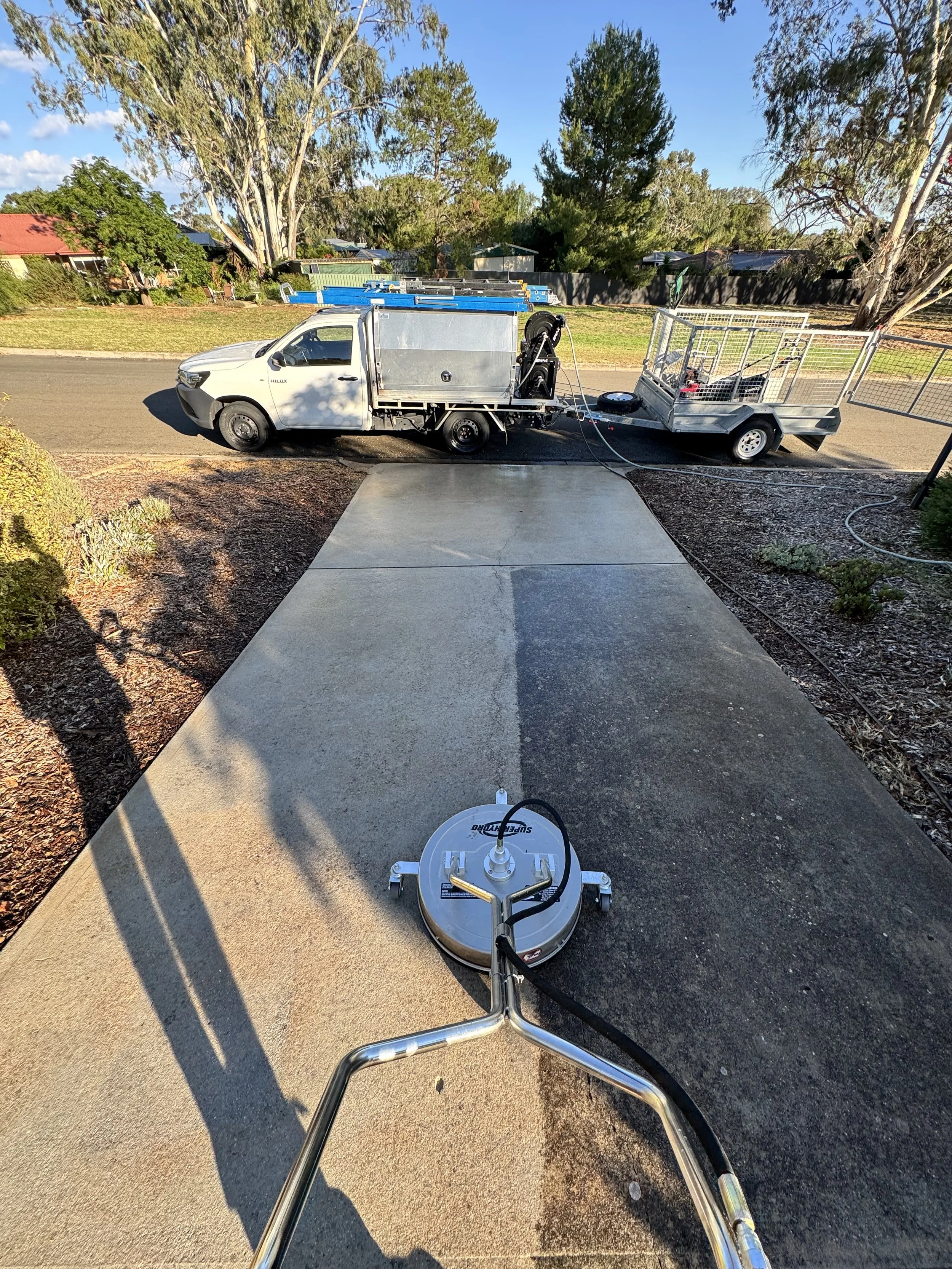 View from a sidewalk showing a concrete surface being cleaned with a power scrubber, connected to a white truck with equipment and a trailer, in a suburban neighborhood with trees and houses in the background.