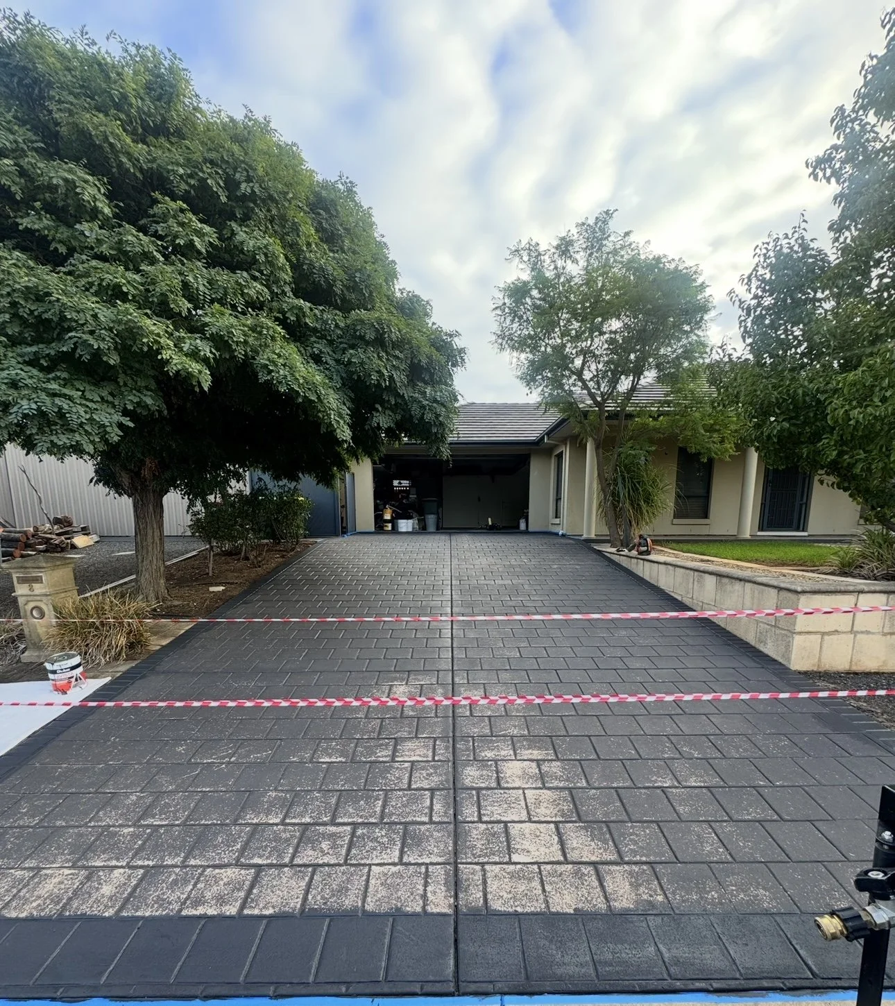 Newly paved asphalt driveway in front of a house, bordered by a stone wall on the right and a large tree on the left, with a partly open garage and a cloudy sky overhead.