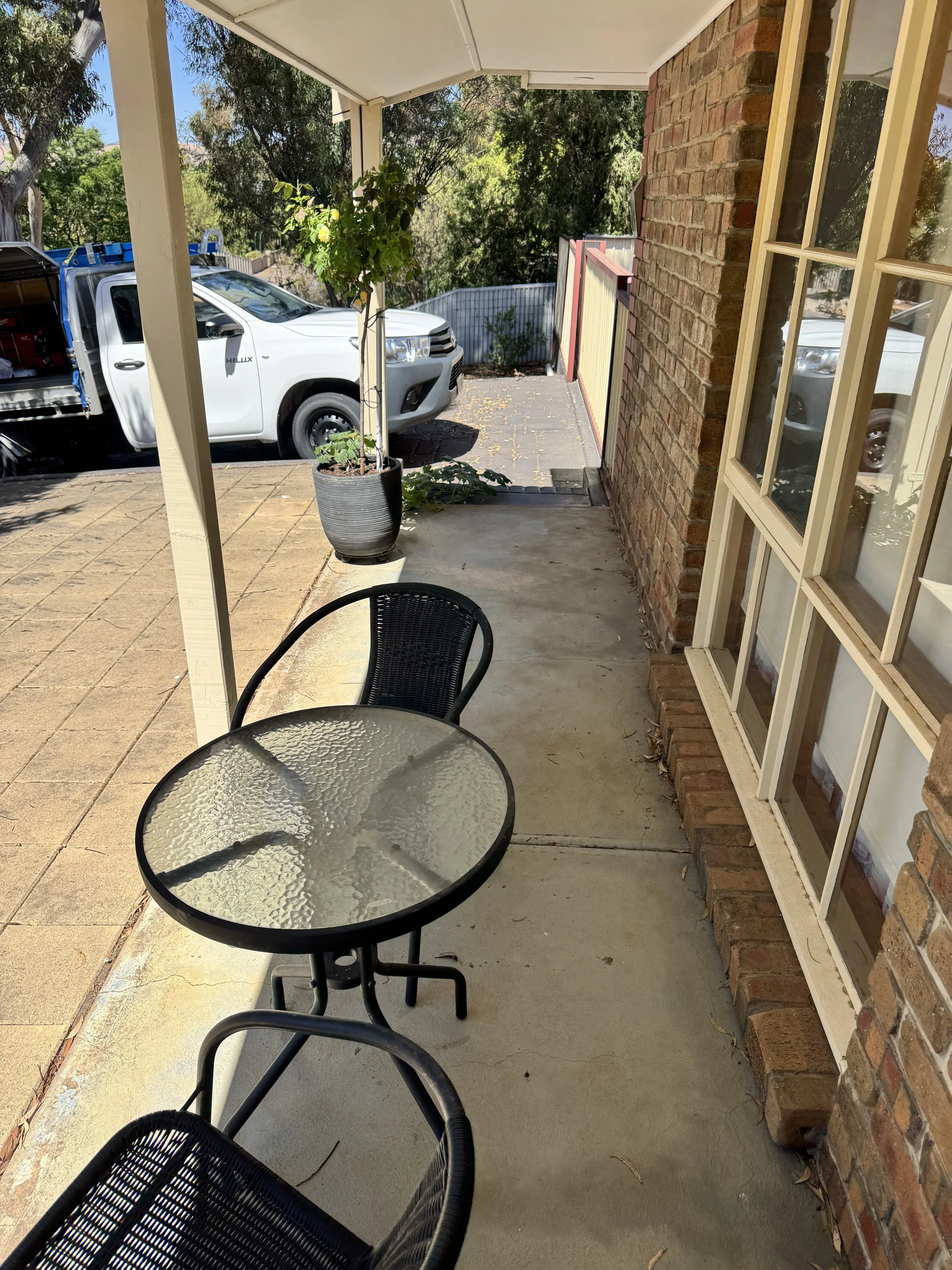 View from a porch showing a glass outdoor table with a black metal chair, a potted plant, a brick house exterior wall, and a driveway with a white truck and trees in the background.