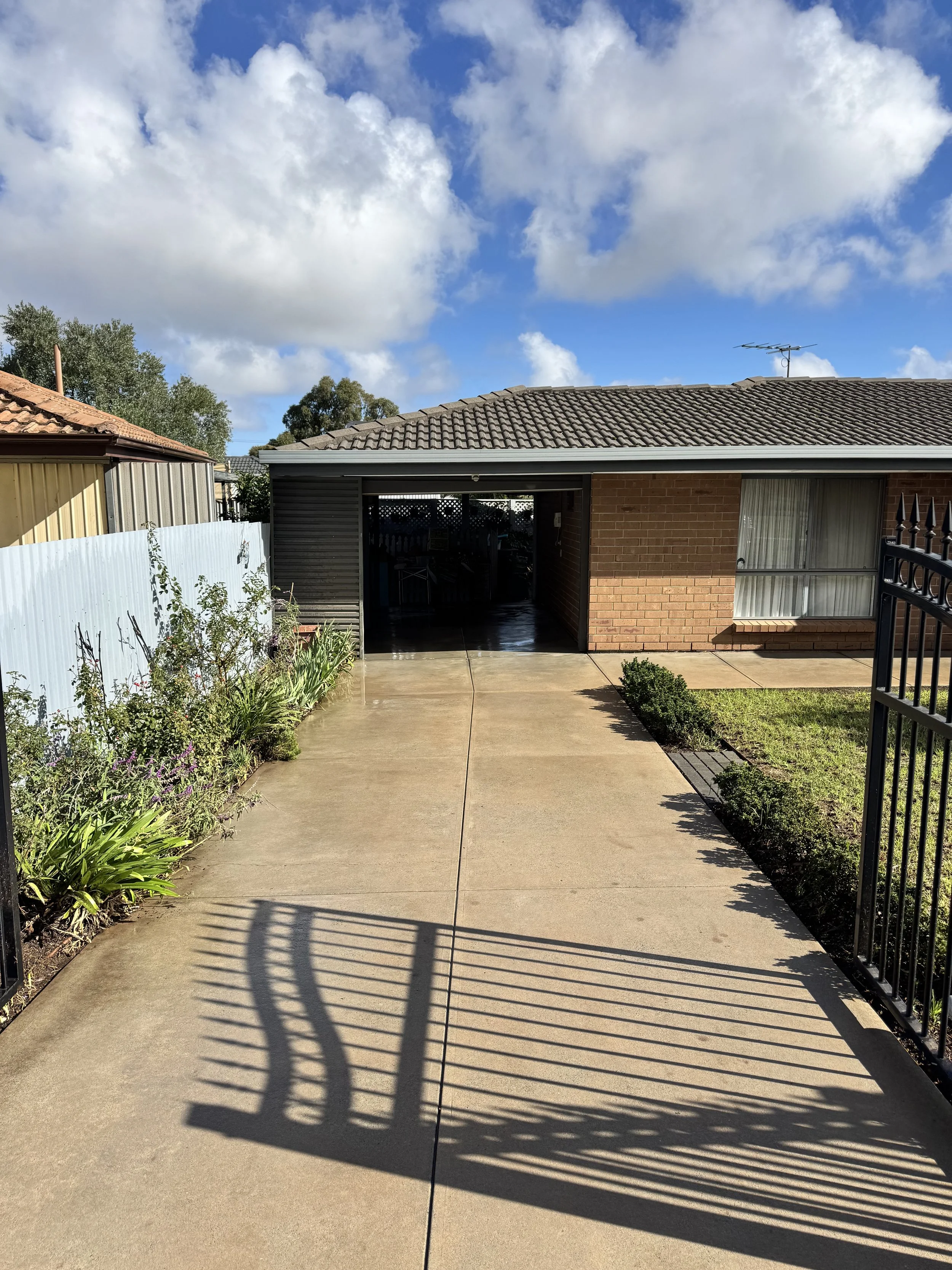 The backyard view shows a concrete patio, garden with various plants, a brick house with a large window, and a black fence on the right side. The sky is partly cloudy with blue and white clouds.
